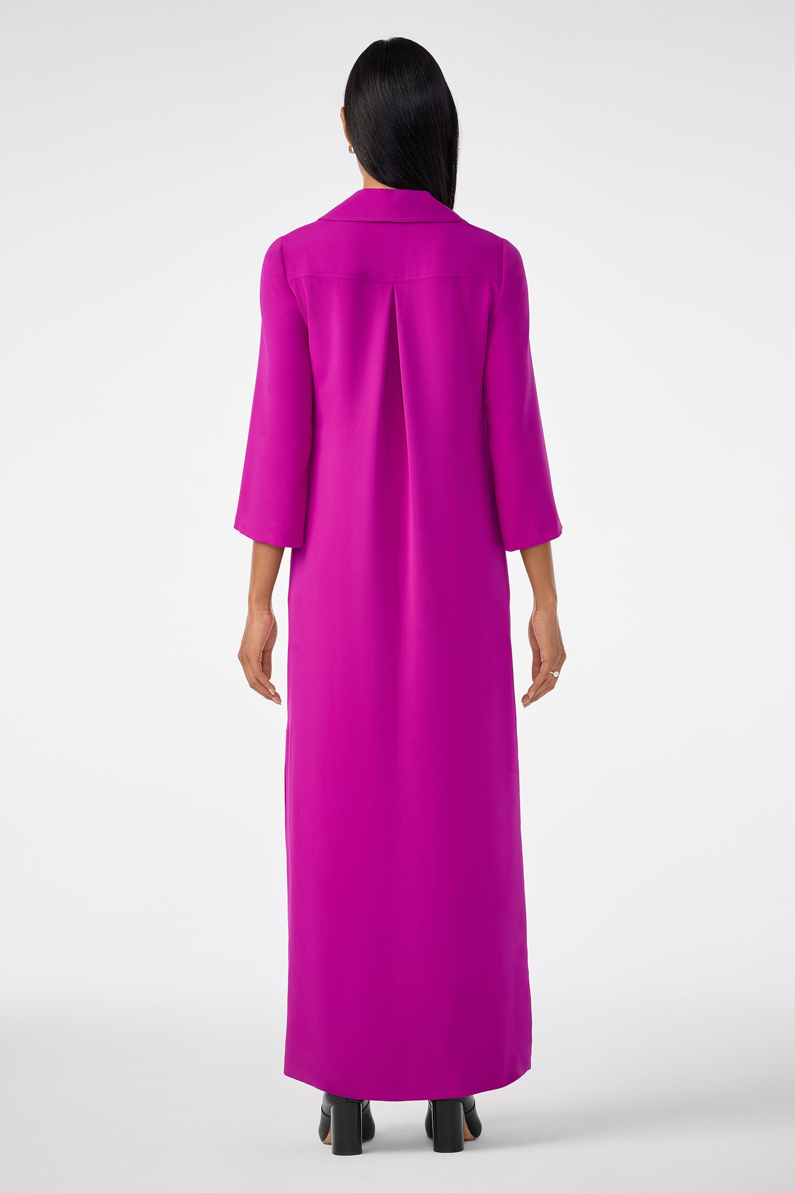 A person stands facing away in the Annecy Dress, a long, vibrant magenta piece with three-quarter sleeves, paired with black heeled shoes against a plain white background.
