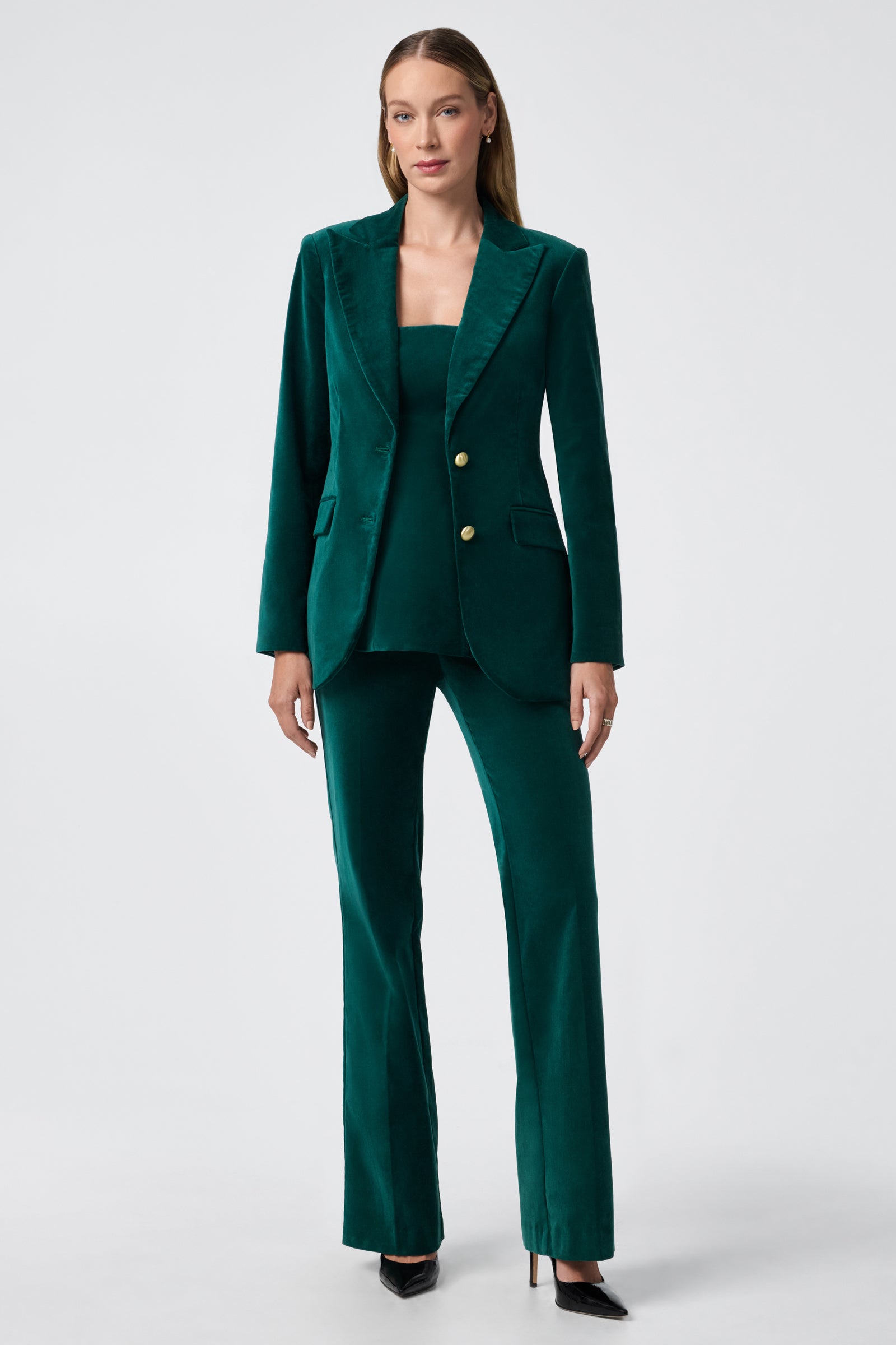 A woman stands against a plain background wearing the Finale Top in deep green velvet, paired with matching trousers and black pointed-toe heels. Her long, straight hair frames her face as she poses with a neutral expression.