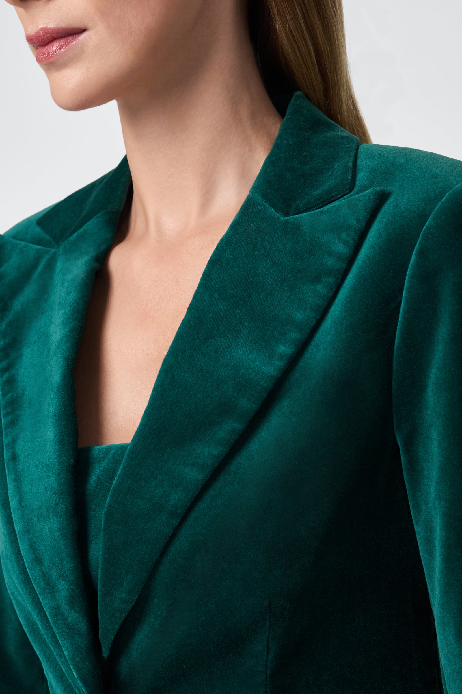 A woman, shown from the shoulders up, wears the deep green Broadway Jacket with a notched lapel. The plain light background highlights the velvet texture and rich color of the blazer.