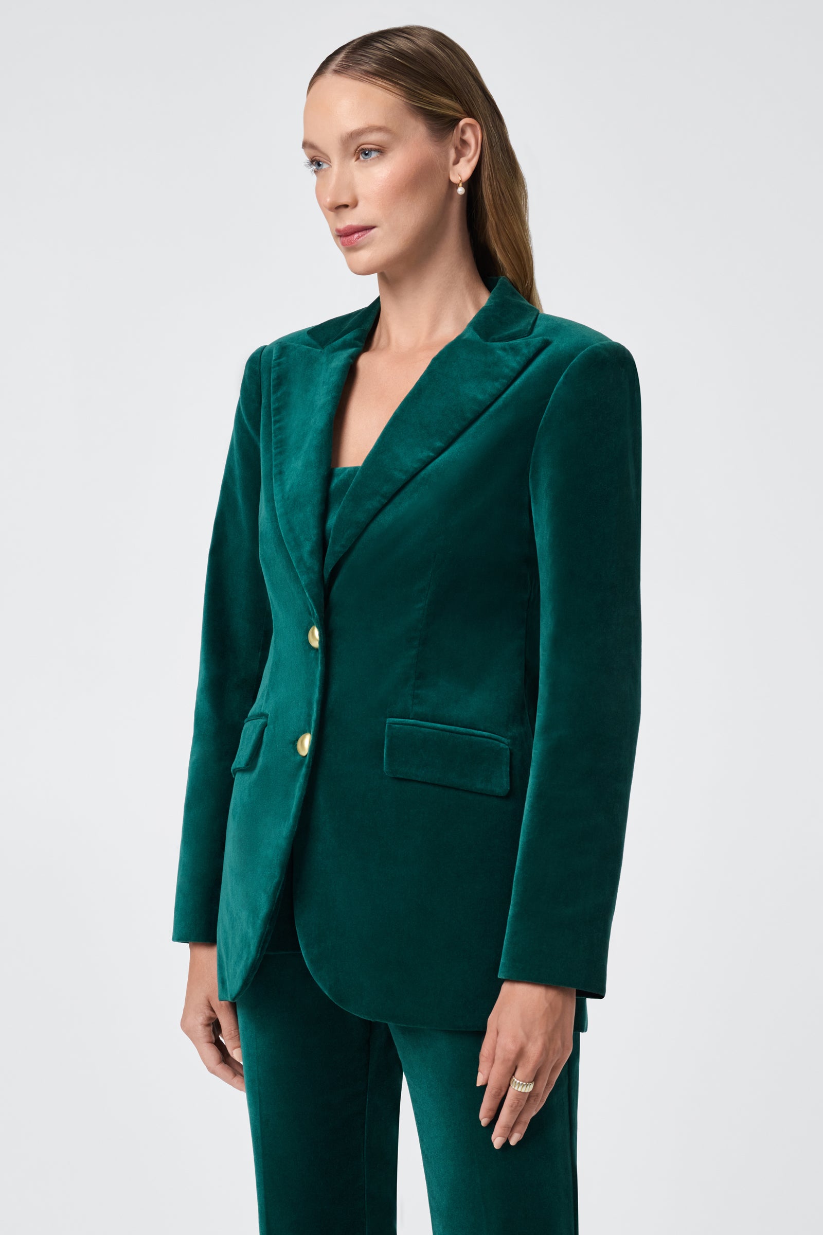 A woman with long, straight hair wears the Broadway Jacket—a dark green velvet blazer with gold buttons—paired with matching pants. She stands against a plain light background, looking slightly to the side.