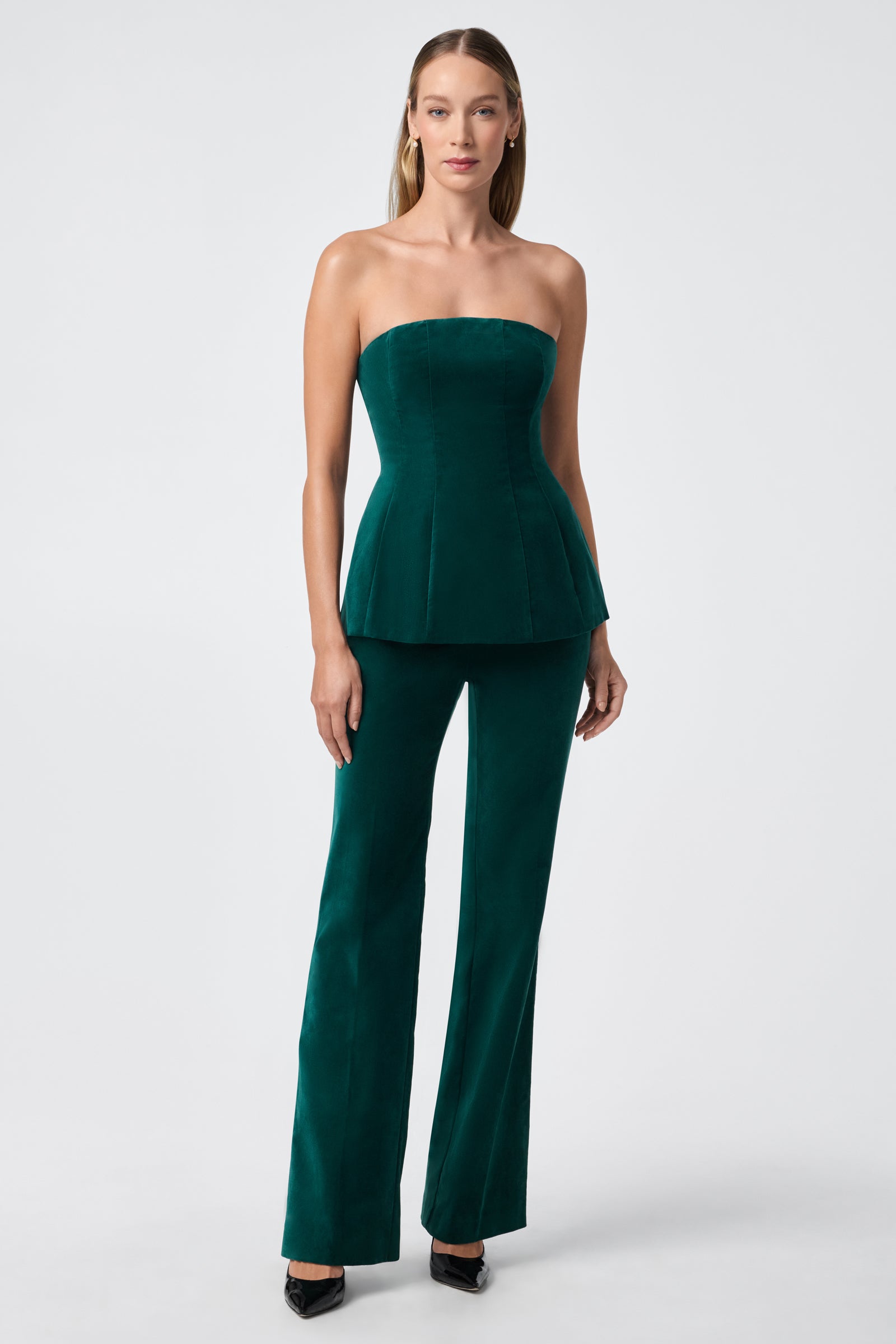 A woman wearing the strapless, fitted dark green velvet Finale Top with matching flared pants and black pointed-toe heels stands against a plain light background.