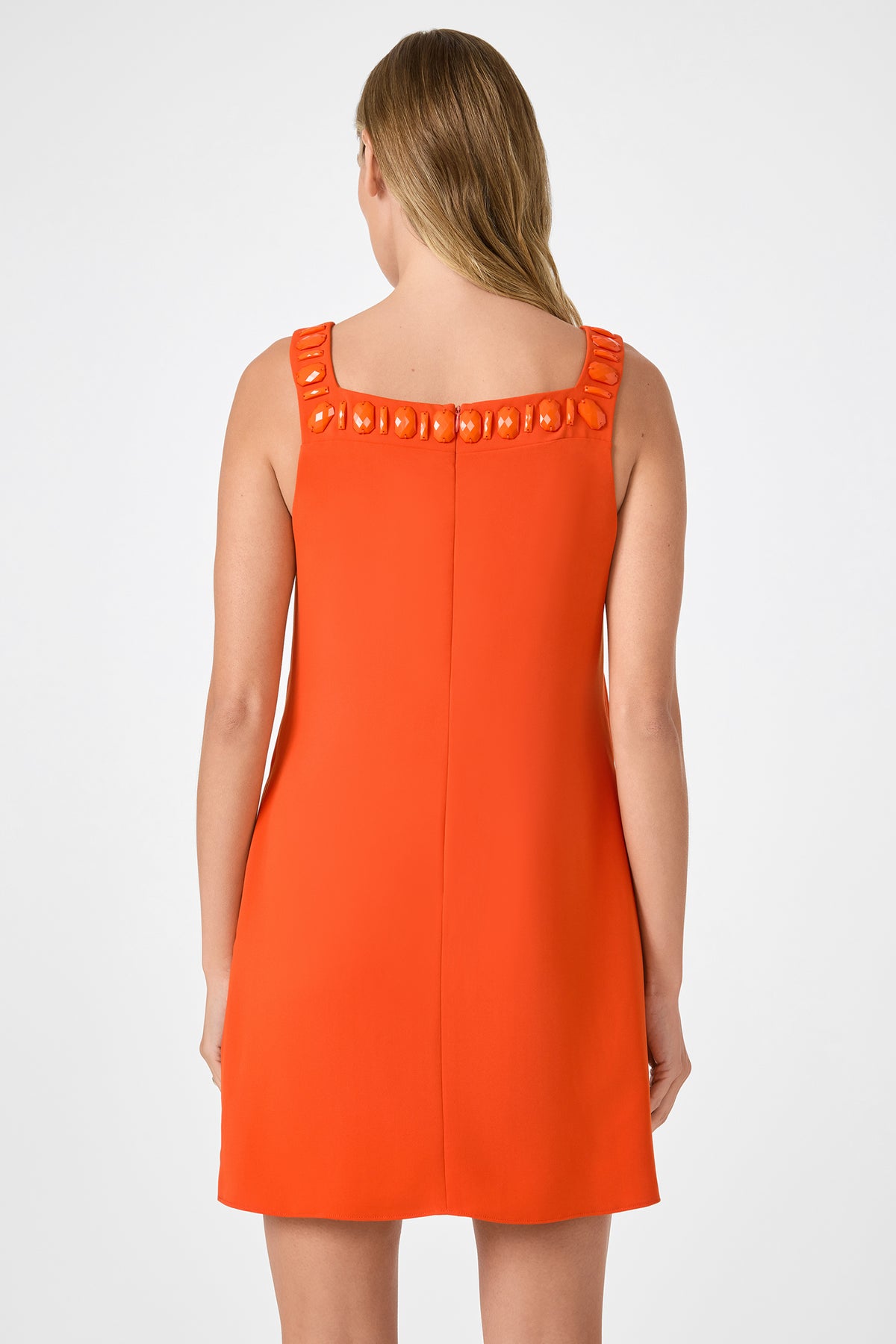 A woman with light brown hair models the Yokuna Jewel Neck Mini Dress, an orange sleeveless style with decorative collar details, shown from the back against a plain white background.
