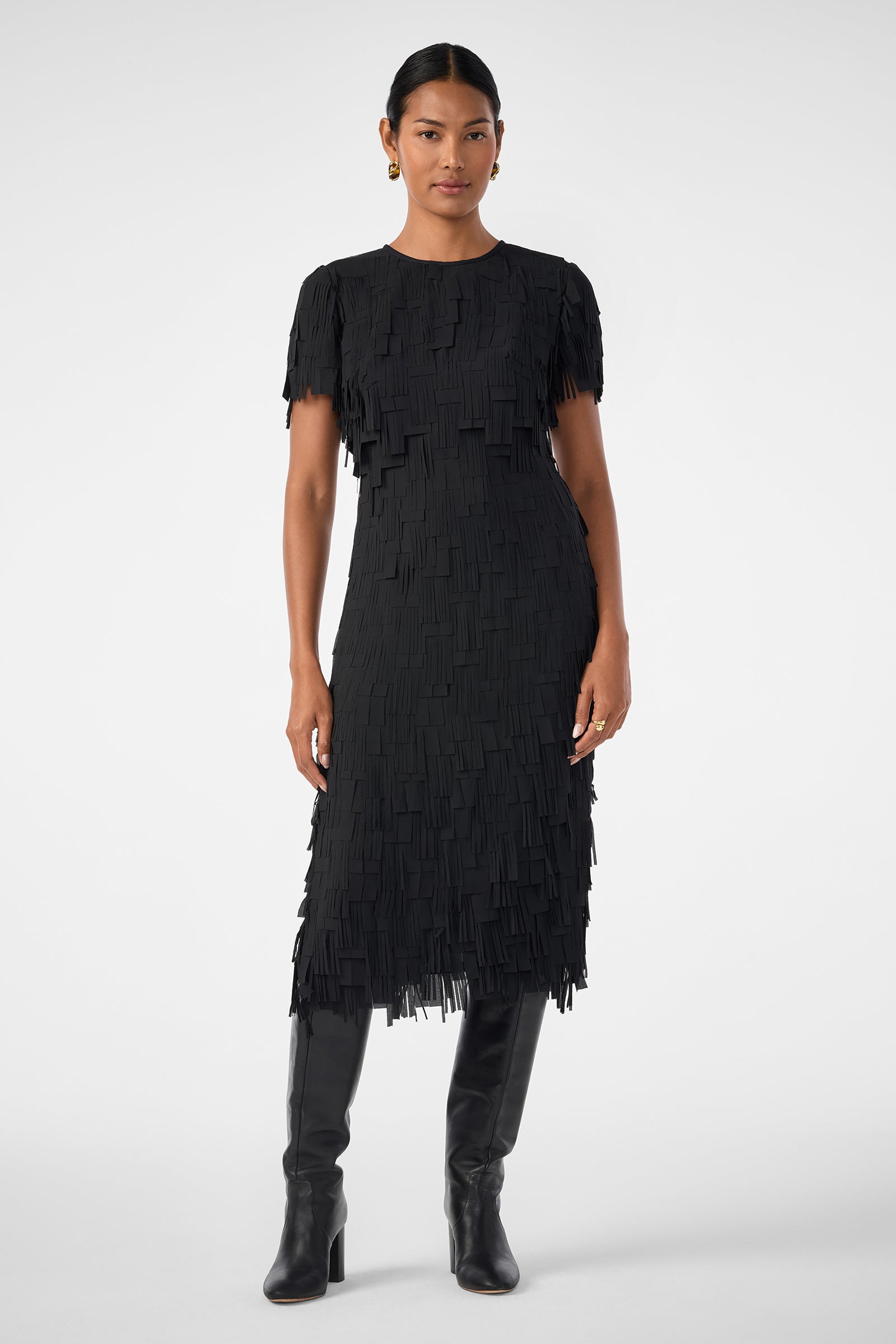 A person stands against a plain background wearing the Silkoak Dress, a black, short-sleeved, knee-length dress with fringe details, paired with tall black boots.