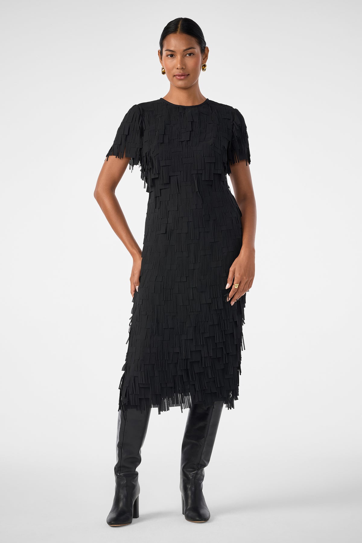 A woman stands confidently with one hand on her hip, wearing the Silkoak Dress—a black, short-sleeved, knee-length dress with a fringed texture—paired with black knee-high boots against a plain white background.