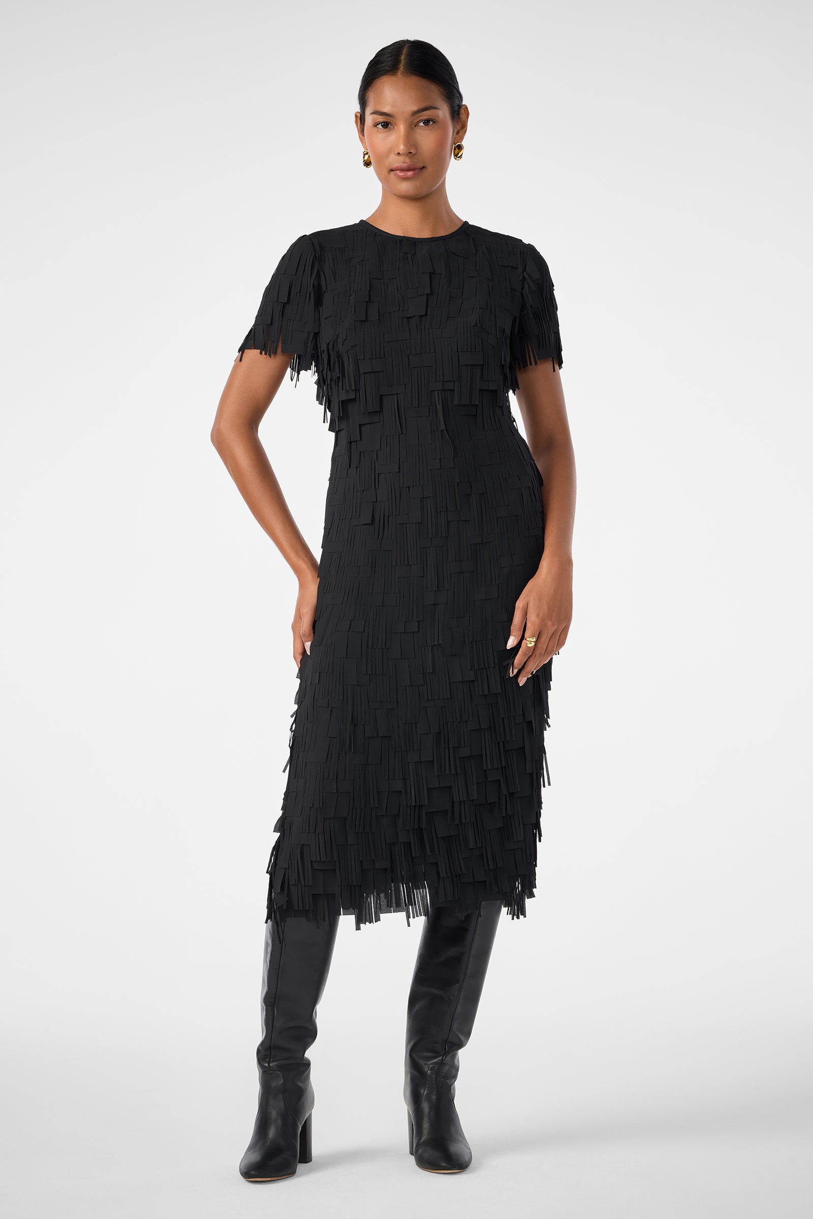 A woman stands confidently with one hand on her hip, wearing the Silkoak Dress—a black, short-sleeved, knee-length dress with a fringed texture—paired with black knee-high boots against a plain white background.