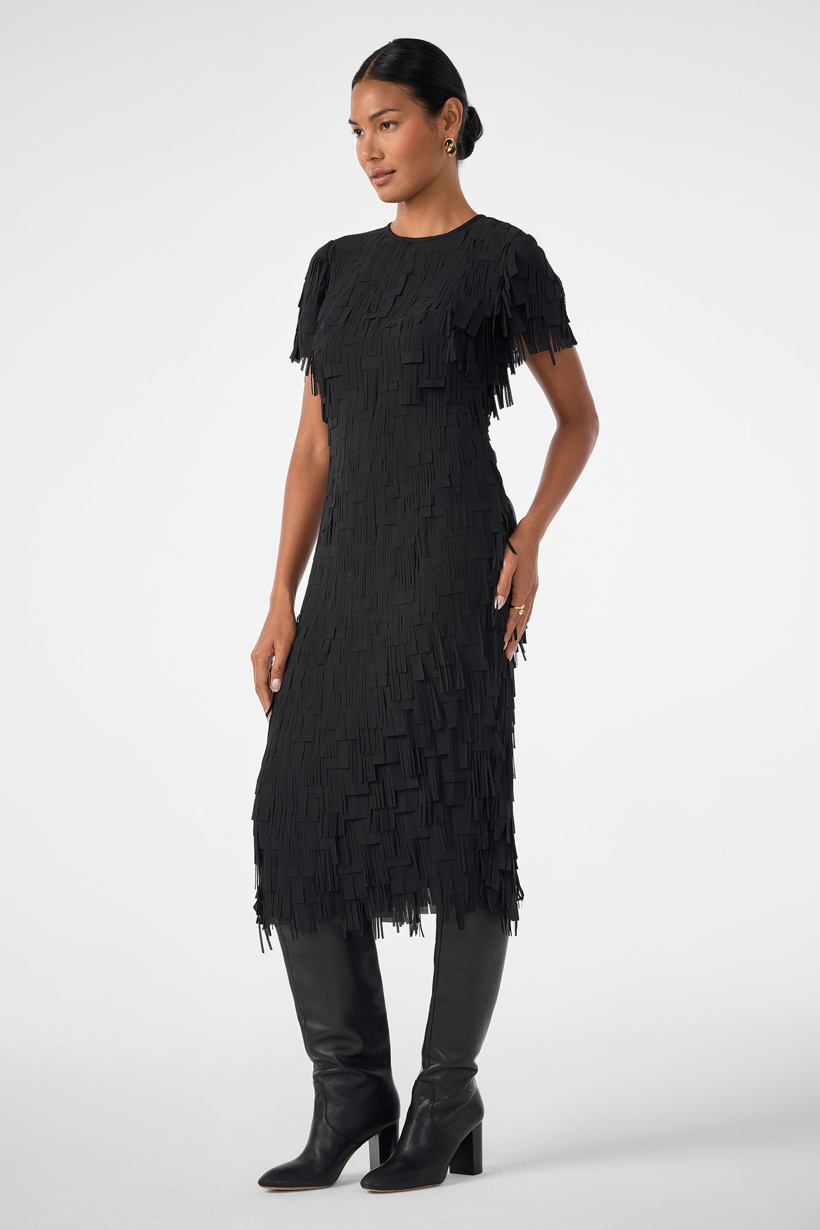 A woman poses against a plain light background, wearing the Silkoak Dress—a textured black midi dress with short sleeves and a layered, fringe-like pattern—paired with black knee-high boots and her hair pulled back.