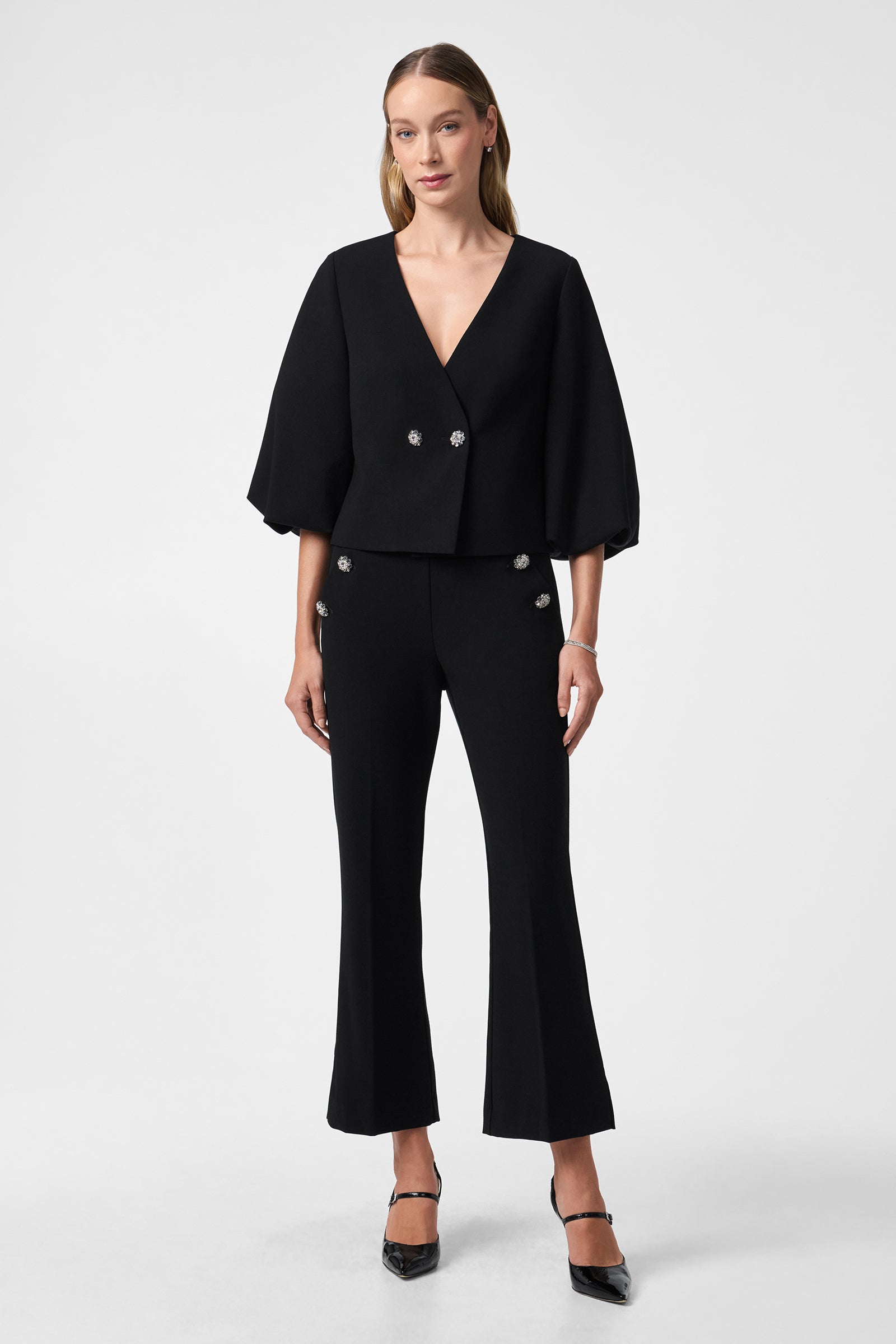 A woman poses in the Basal Cropped Jacket, a stylish black V-neck top with three-quarter sleeves, large decorative buttons, and matching flared pants. She completes the look with black heels against a plain background.