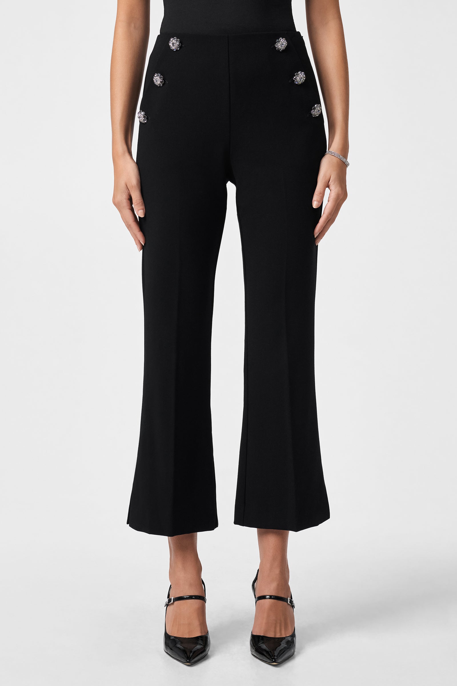 A woman wearing the Luhrmann Pant—high-waisted, black cropped pants with silver front buttons—paired with black pointed-toe heels, stands against a plain white background.