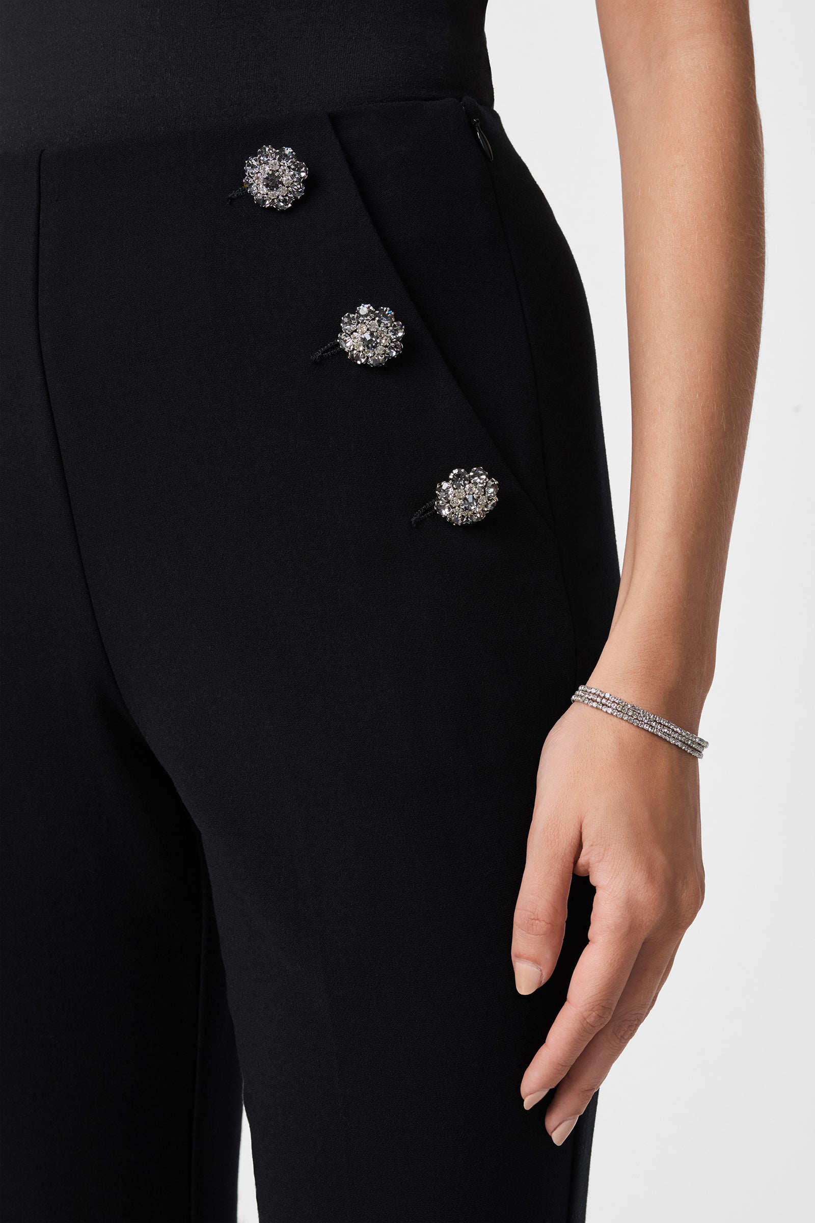 Wearing the Luhrmann Pant, which features four decorative, flower-shaped side embellishments, and a silver bracelet. Only the lower torso and right arm are visible.