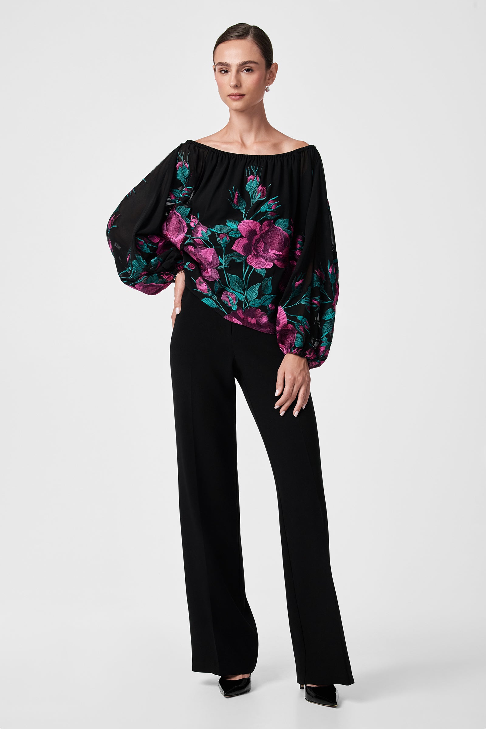 A woman wears the Soho 2 Top—a black off-the-shoulder blouse with voluminous sleeves and a bold pink floral print—paired with high-waisted, wide-leg black pants and heels, posing against a white background.