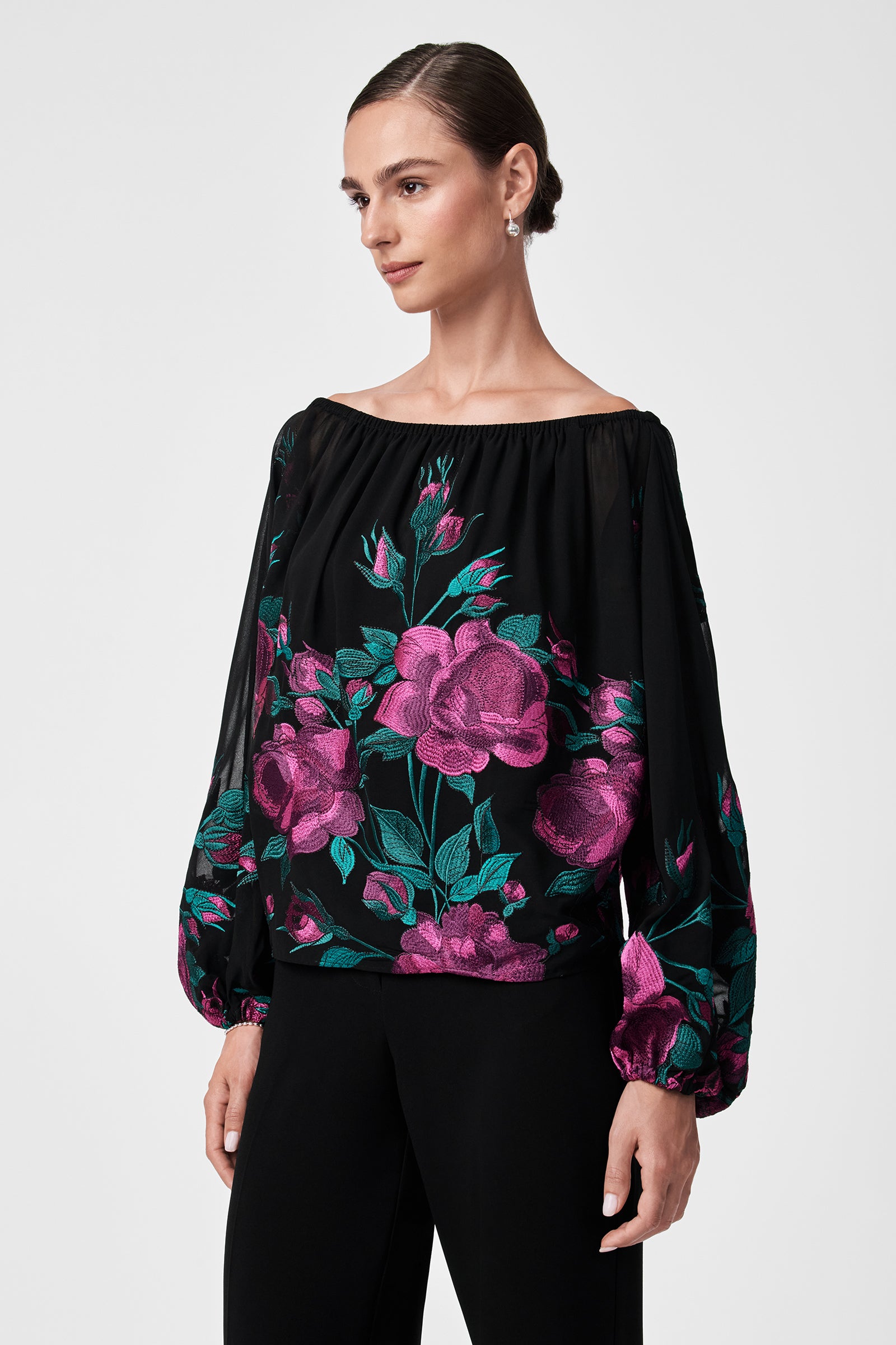 A woman with dark hair pulled back is wearing the Soho 2 Top, a black off-the-shoulder blouse featuring long sleeves and a vibrant pink and teal floral pattern. She stands against a plain light background, facing slightly to the side.