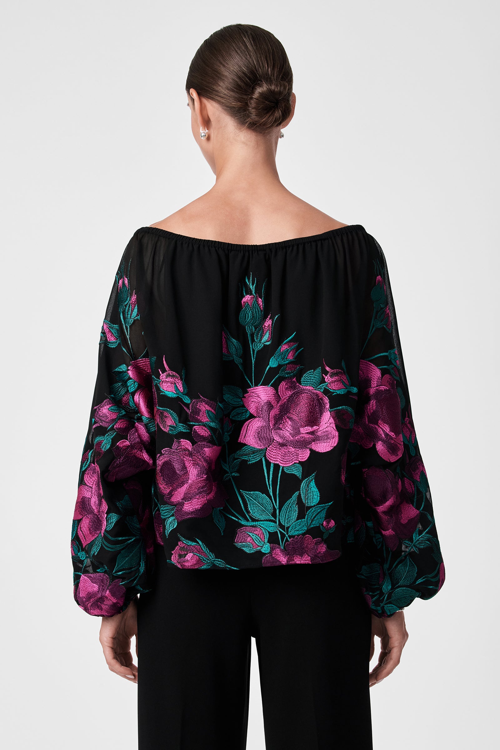 A woman with hair in a low bun is seen from behind, wearing the Soho 2 Top—an off-the-shoulder black blouse with voluminous sleeves and a bold pink and green floral pattern—paired with black pants.