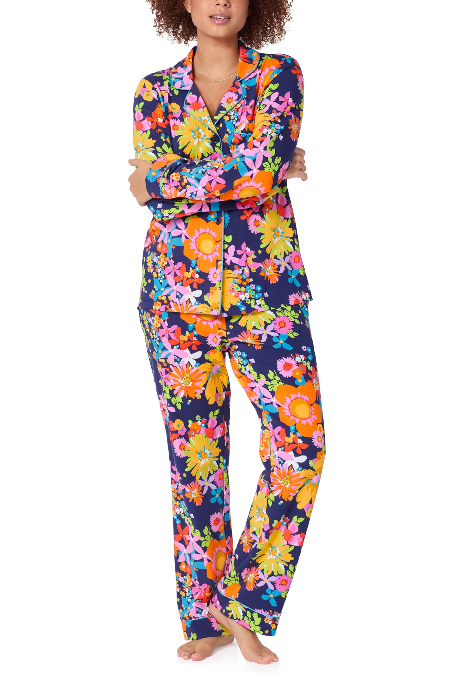 A person stands barefoot with arms crossed, wearing the Classic PJ Set - Bay St Bloom: a long-sleeve, button-up pajama set featuring a vibrant floral pattern on a dark background.