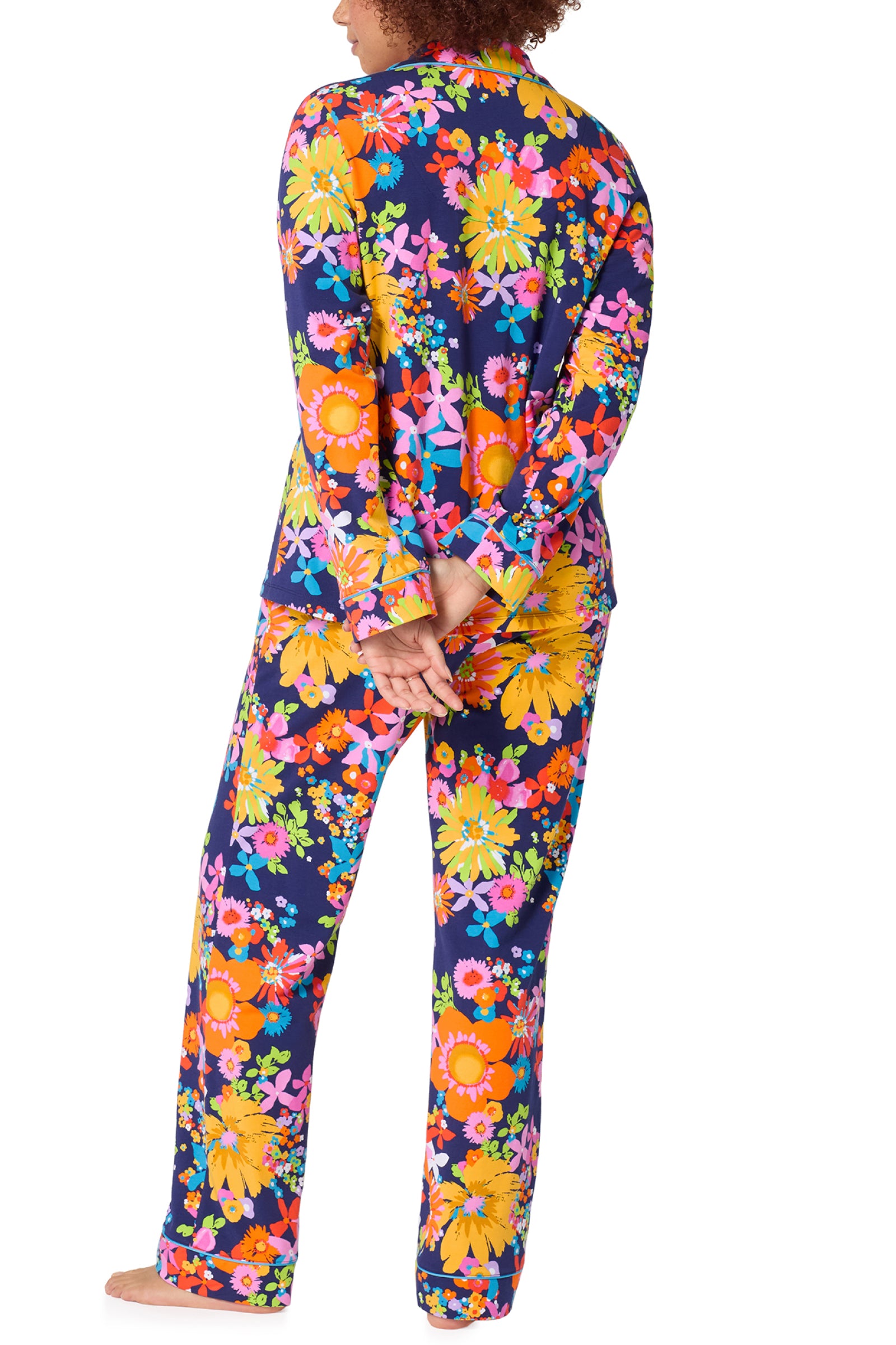 A person stands barefoot with hands behind their back, wearing the Classic PJ Set - Bay St Bloom, a matching long-sleeve top and pants with a vibrant floral pattern on a dark background, seen from behind.