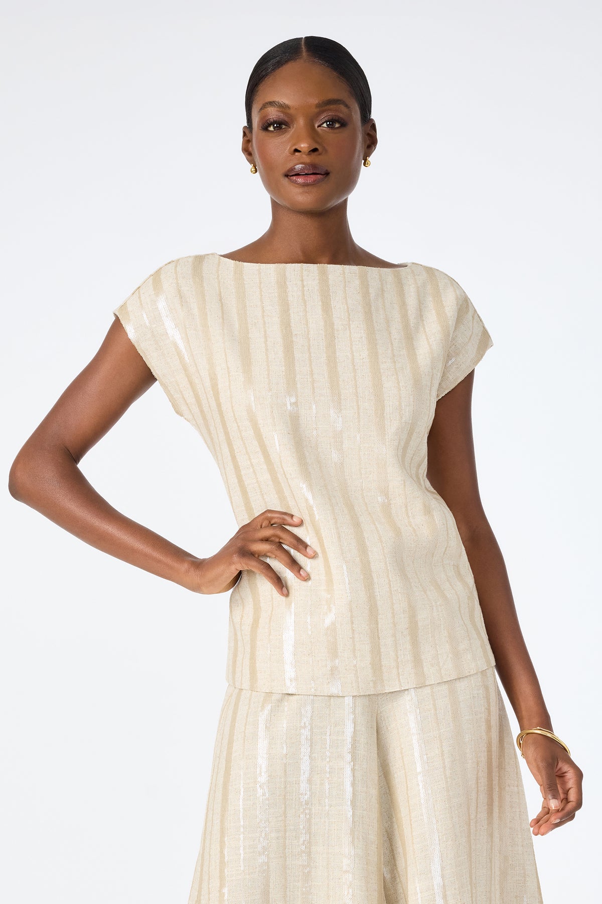 A woman poses against a white background wearing the Breene Boatneck Top, a sleeveless, beige, shimmery piece paired with a matching skirt. She accessorizes with small hoop earrings and a bracelet, her short hair pulled back.