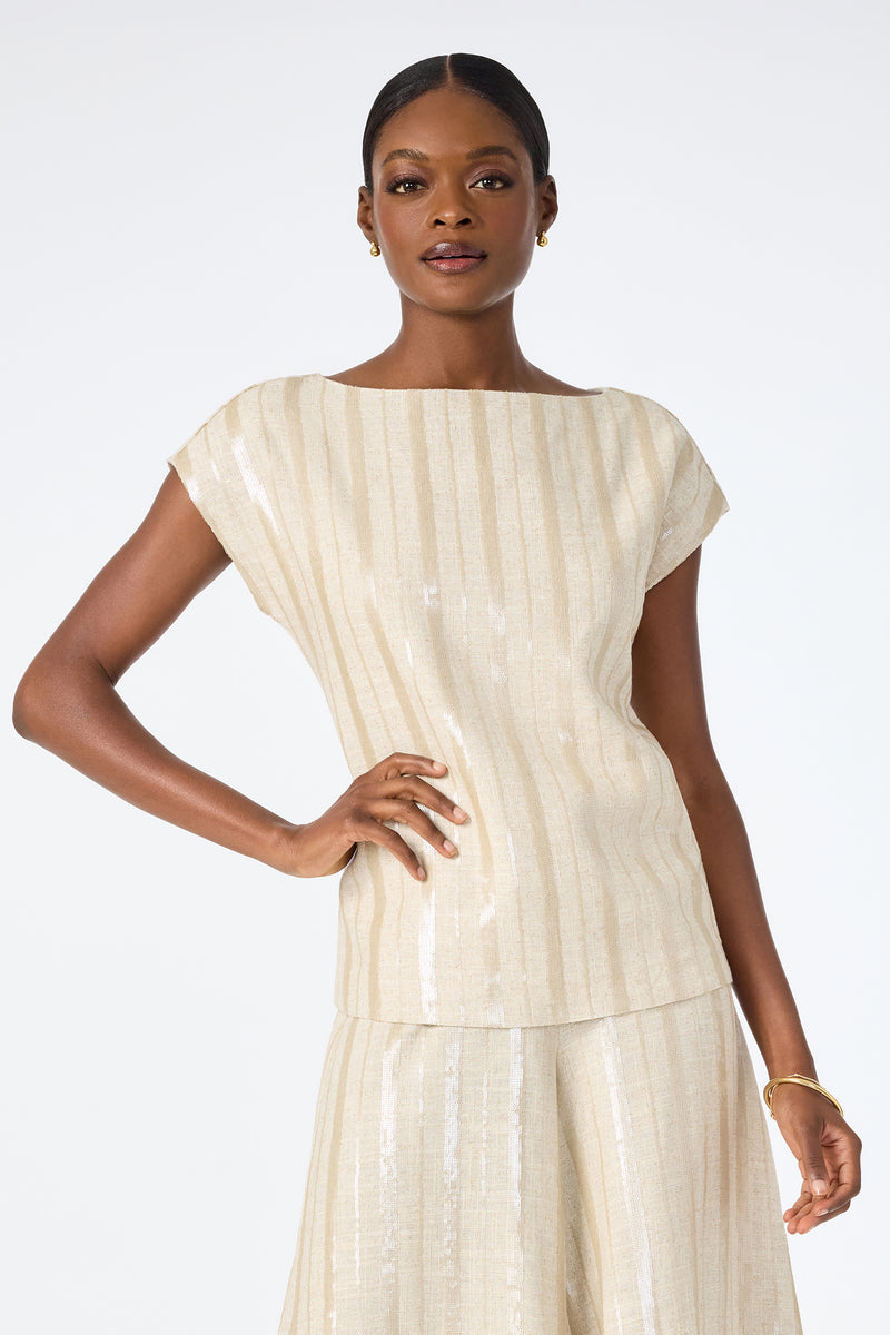 A woman poses against a white background wearing the Breene Boatneck Top, a sleeveless, beige, shimmery piece paired with a matching skirt. She accessorizes with small hoop earrings and a bracelet, her short hair pulled back.