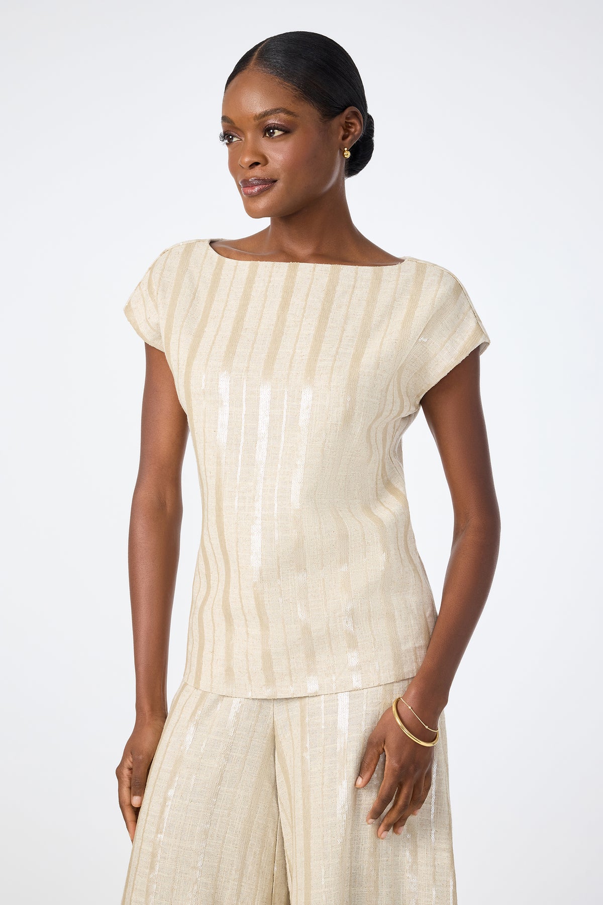 A woman wears the Breene Boatneck Top‚Äö√Ñ√Æa light beige, short-sleeve, striped blouse‚Äö√Ñ√Æpaired with matching wide-leg pants, standing against a plain white background and smiling gently as she looks to the side.