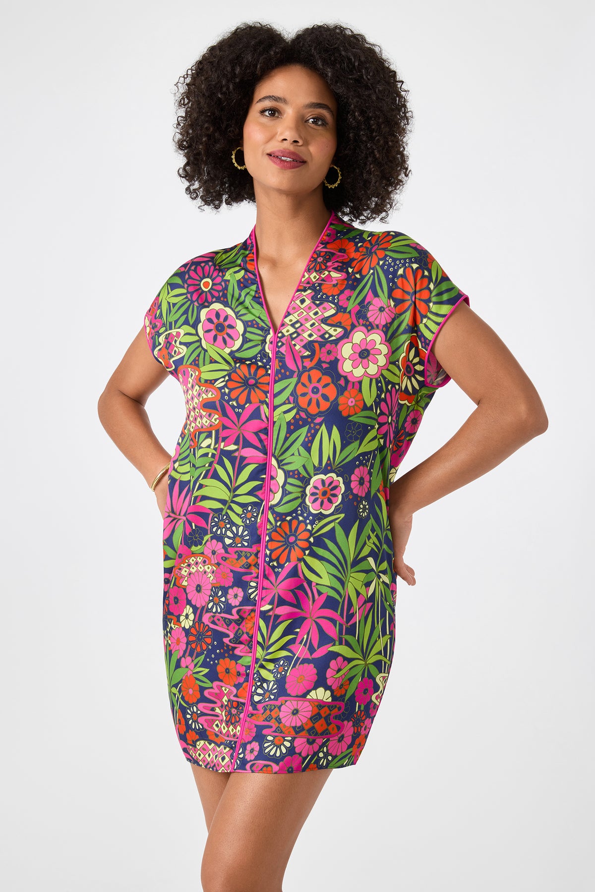 A woman stands with hands in pockets against a light background, wearing the Tarashi Short-Sleeve Mini Dress - Fumi Floral, featuring a vibrant multicolored floral print on a dark base and a comfortable mini-length fit.