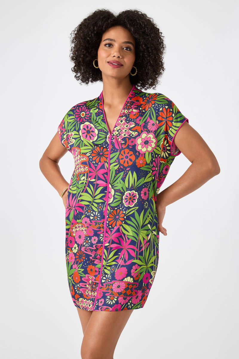 A woman stands with hands in pockets against a light background, wearing the Tarashi Short-Sleeve Mini Dress - Fumi Floral, featuring a vibrant multicolored floral print on a dark base and a comfortable mini-length fit.