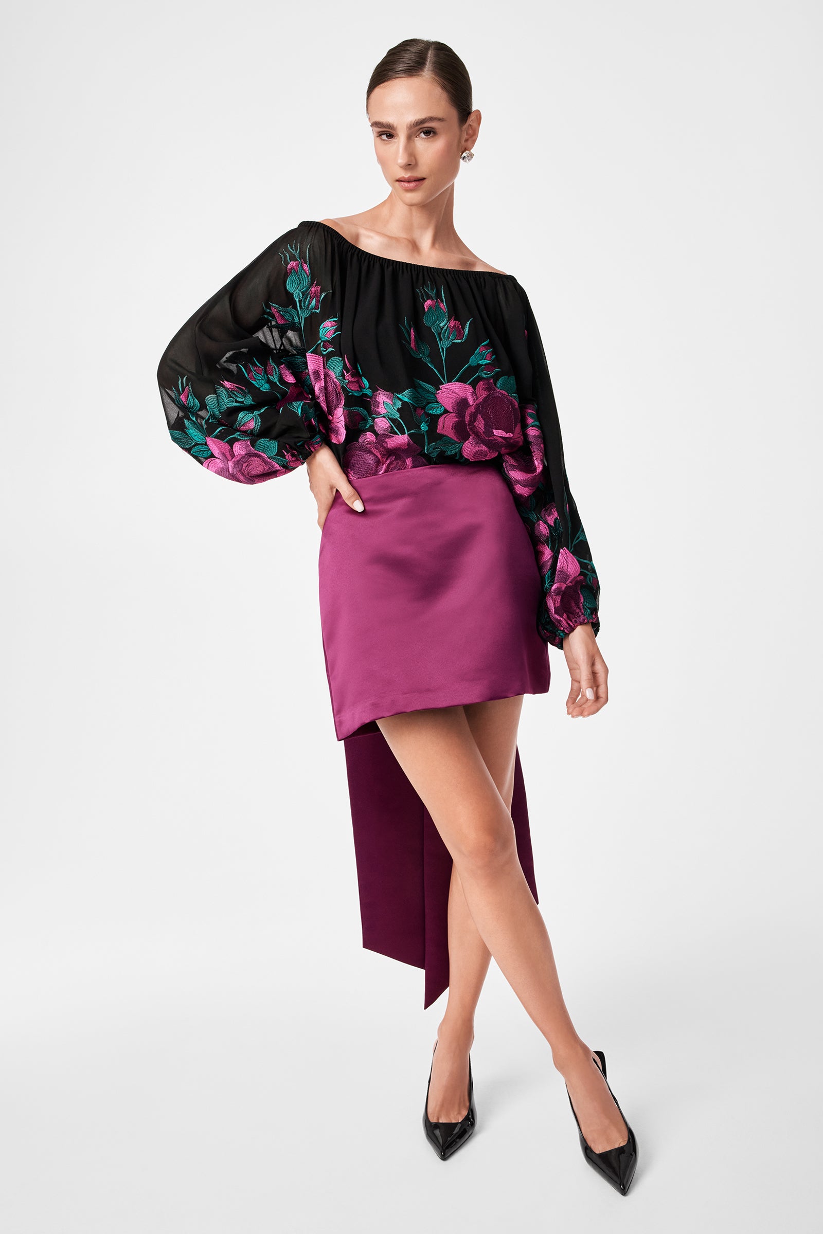 A woman poses in the Soho 2 Top, a black off-the-shoulder blouse with pink floral print and billowy sleeves, styled with a short magenta skirt and black pointed-toe heels against a plain white background.