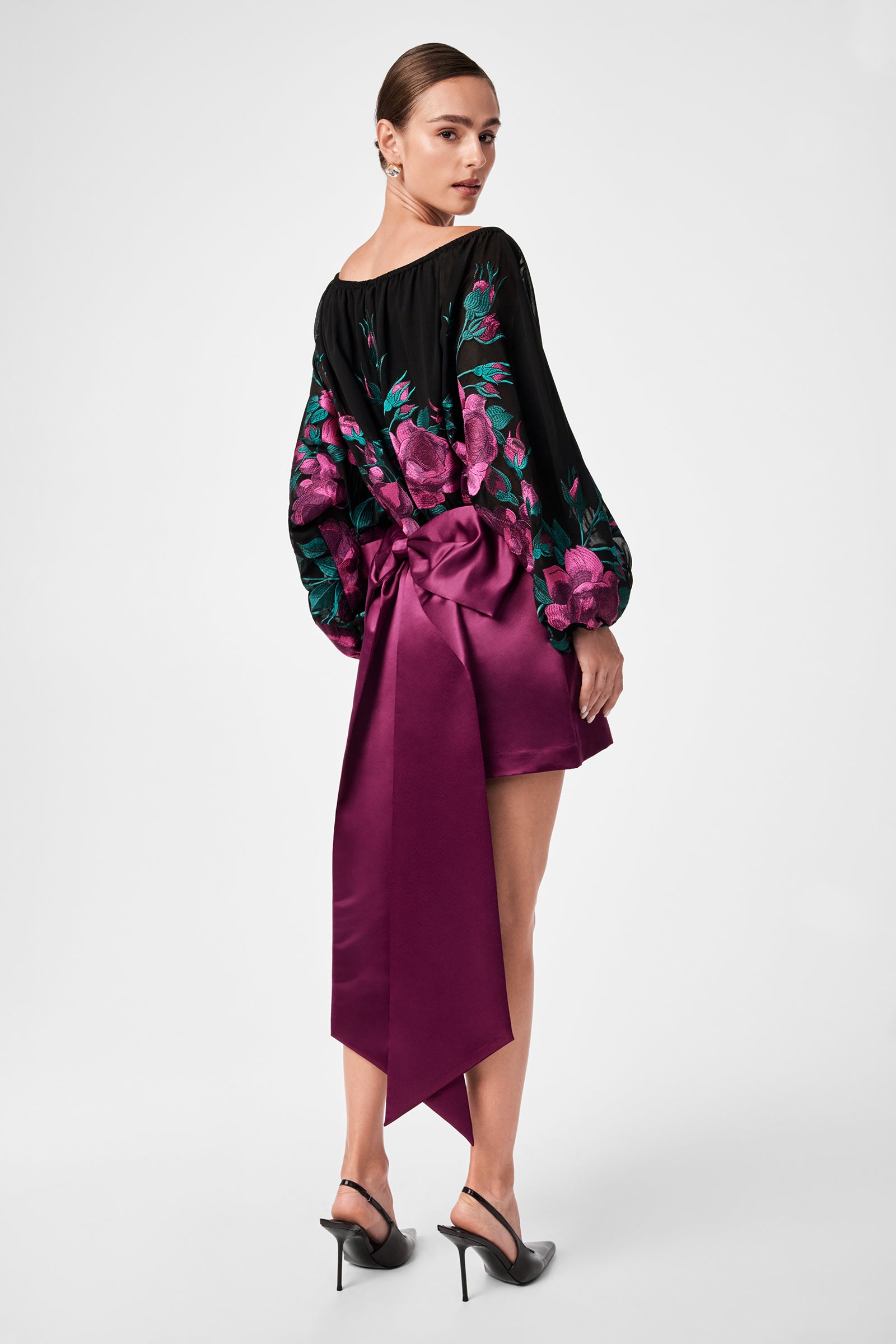A woman faces away, wearing a black top with purple floral patterns and long sleeves, paired with the Vinna Bow Satin Mini Skirt in purple, featuring a large bow and tails. She finishes the look with black heels and her hair tied back.