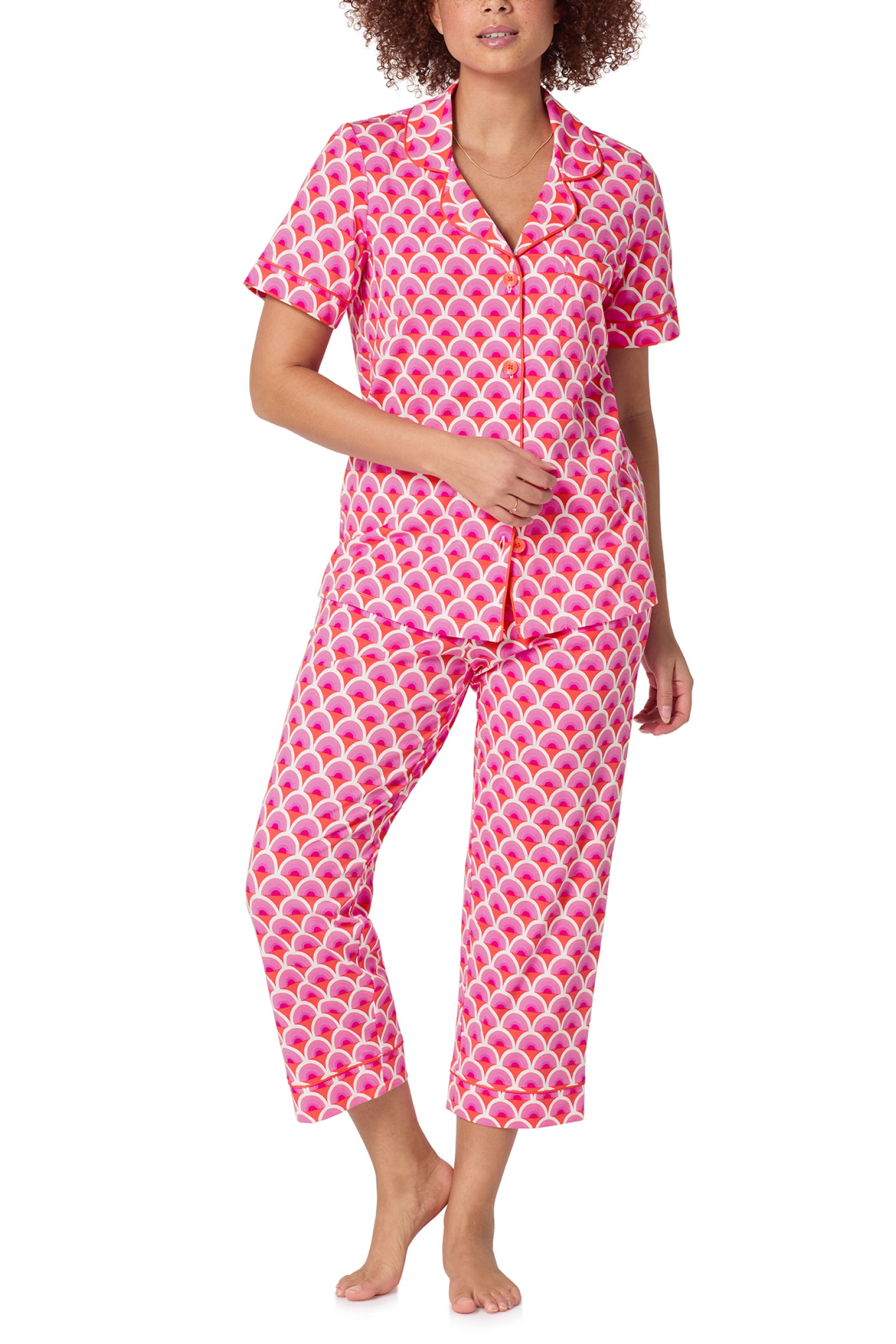 A person stands barefoot wearing the Crop Classic PJ Set - Scallop, a matching pink and white patterned pajama set with short sleeves, cropped pants, a scallop-like design, and front buttons on the shirt.