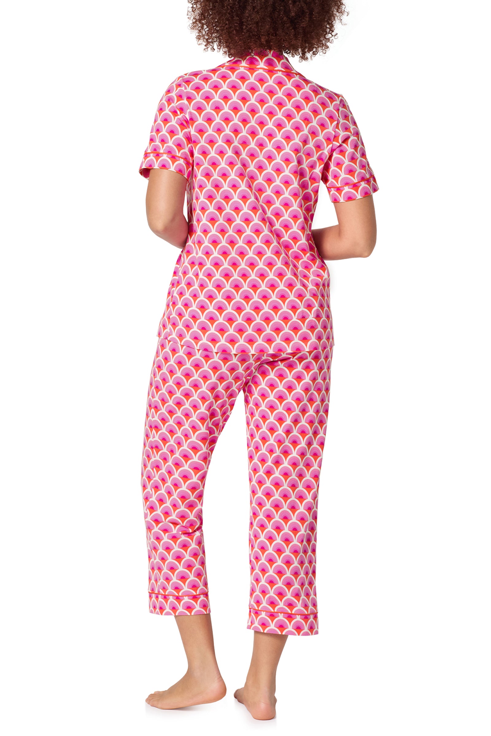 A person stands barefoot facing away, wearing the Crop Classic PJ Set - Scallop, featuring a pink and white pattern with short sleeves and cropped pants.