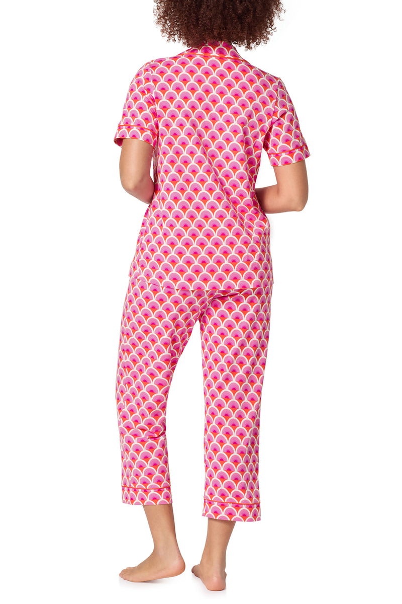 A person stands barefoot facing away, wearing the Crop Classic PJ Set - Scallop, featuring a pink and white pattern with short sleeves and cropped pants.