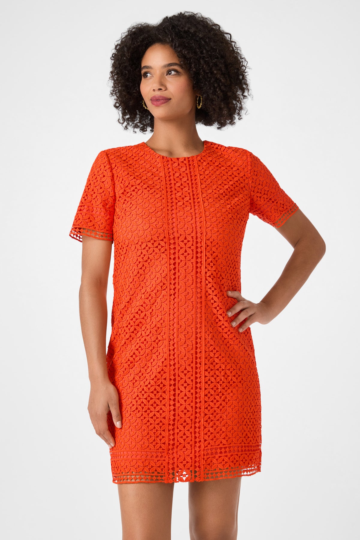 A woman models the Kimasu Short-Sleeve Lace Mini Dress in vibrant orange, standing with one hand on her hip and looking to the side against a plain light background.