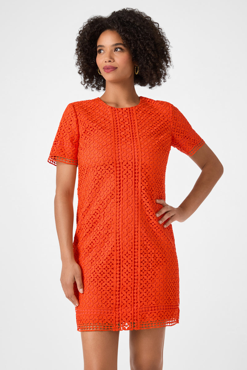 A woman models the Kimasu Short-Sleeve Lace Mini Dress in vibrant orange, standing with one hand on her hip and looking to the side against a plain light background.