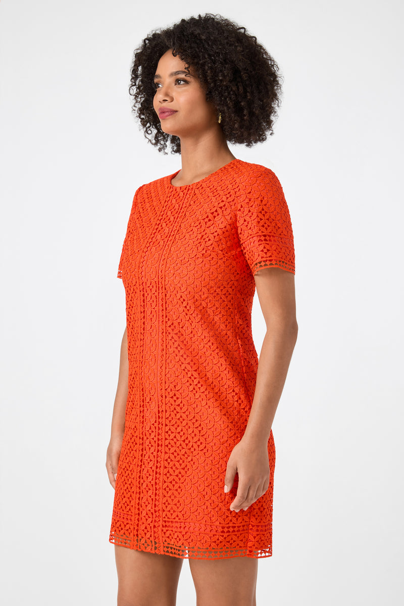 A woman with curly hair wears the Kimasu Short-Sleeve Lace Mini Dress in bright orange, standing against a plain light background and looking slightly to the side.