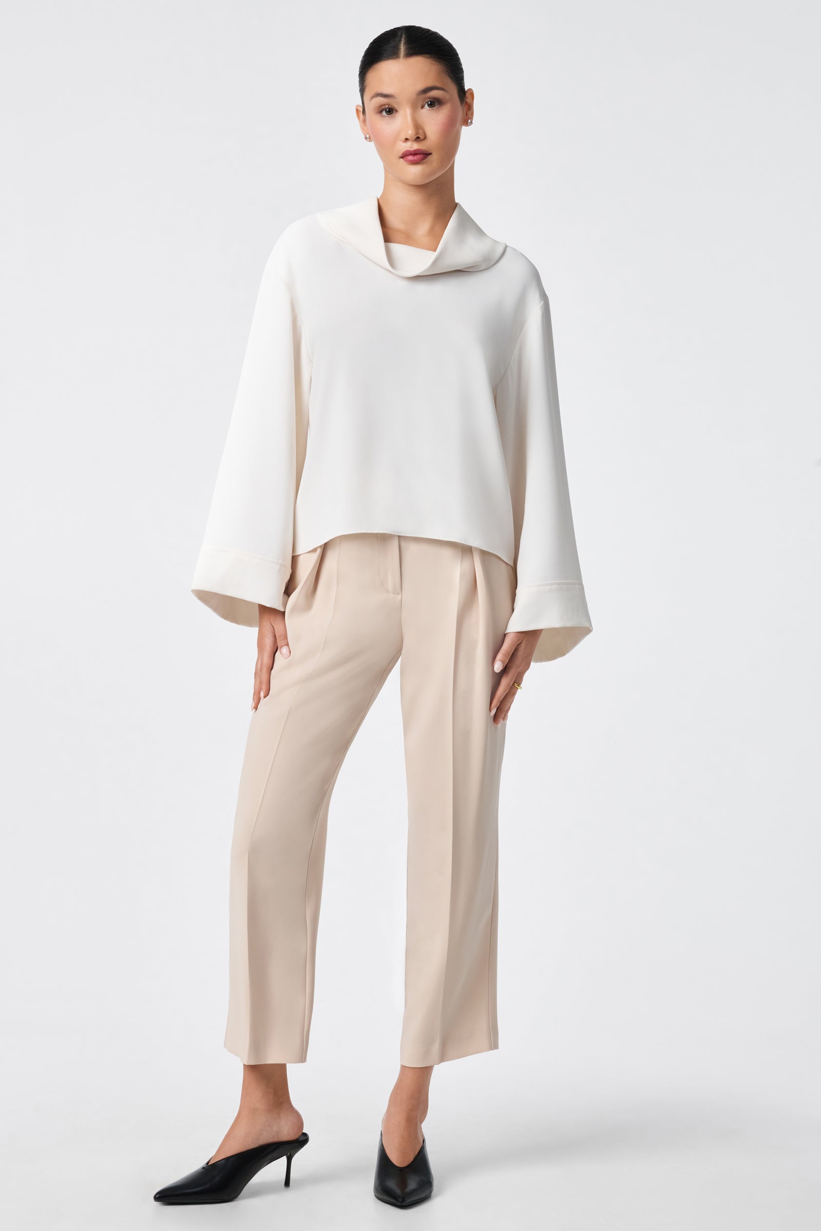 A woman stands against a plain background wearing the Staged Top, a loose white blouse with wide sleeves, styled with beige cropped trousers and black heeled mules. Her hair is pulled back as she poses with one knee bent and a hand on her thigh.