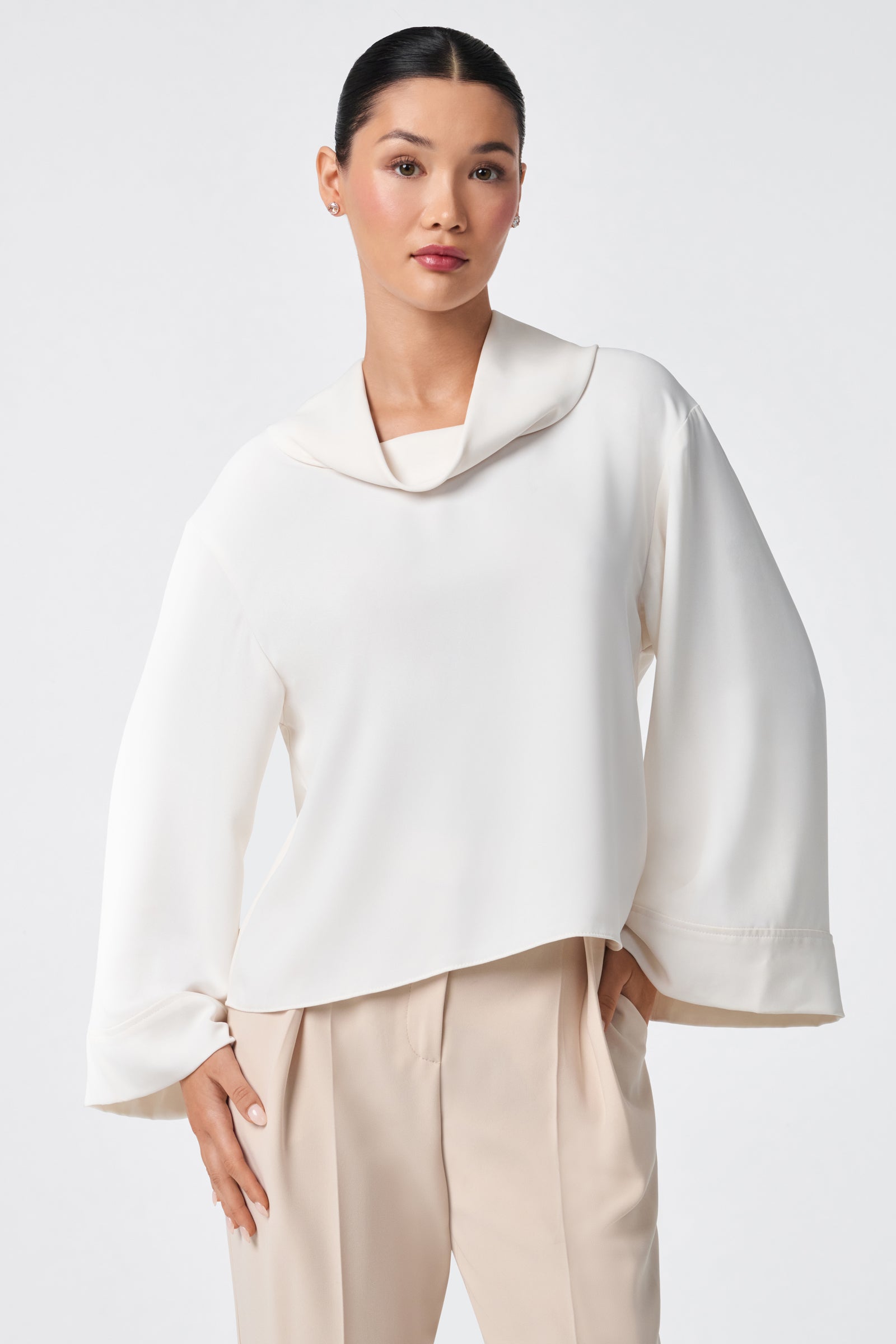 A woman with dark hair in a sleek bun wears the Staged Top, a white, long-sleeve blouse with a wide collar and loose fit, paired with beige pleated pants. She poses with one hand in her pocket against a plain light background.