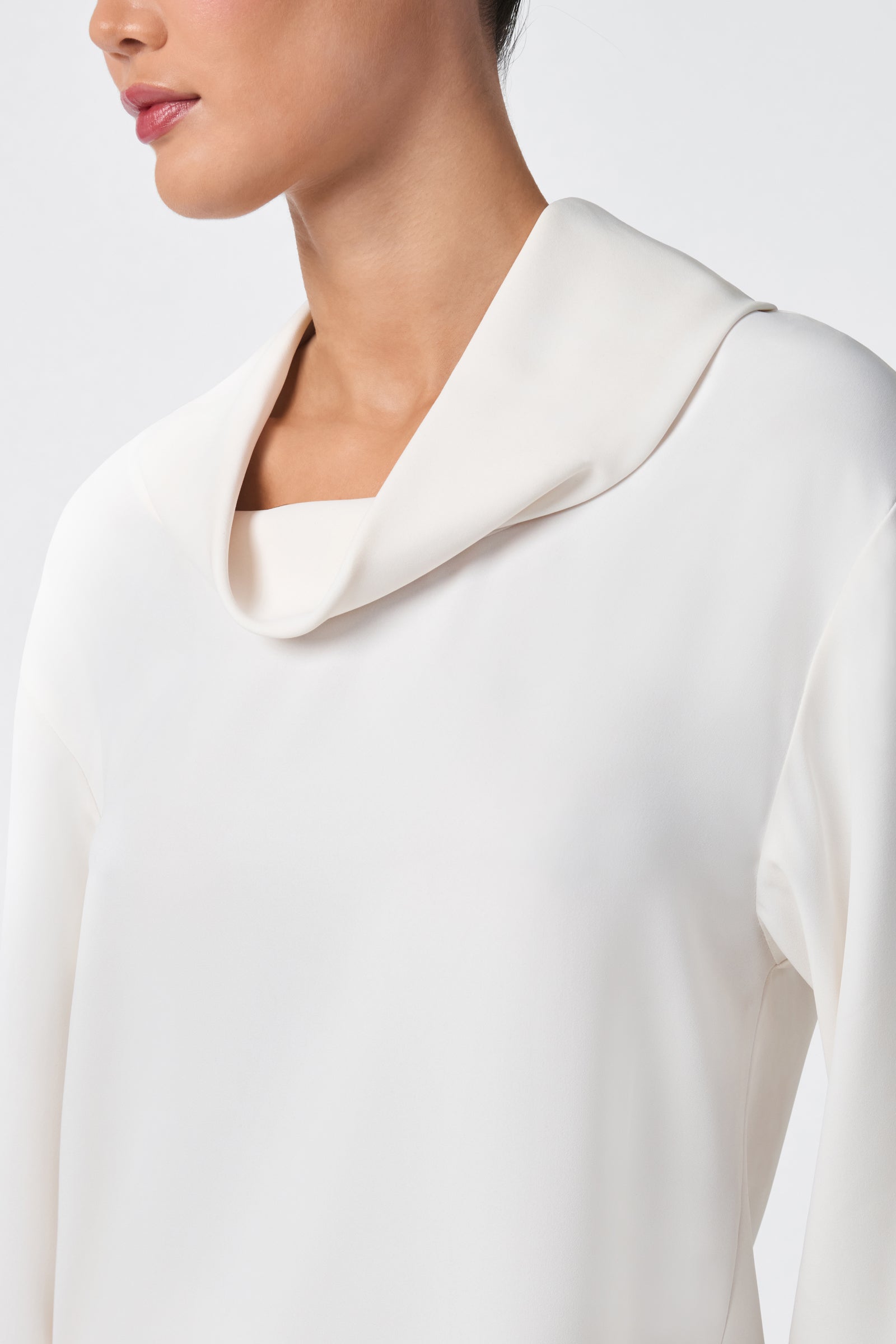 A woman models the Staged Top, an elegant long-sleeved white blouse with a draped cowl neckline. The photo, cropped from nose to waist, highlights its smooth fabric and minimalist style against a light background.
