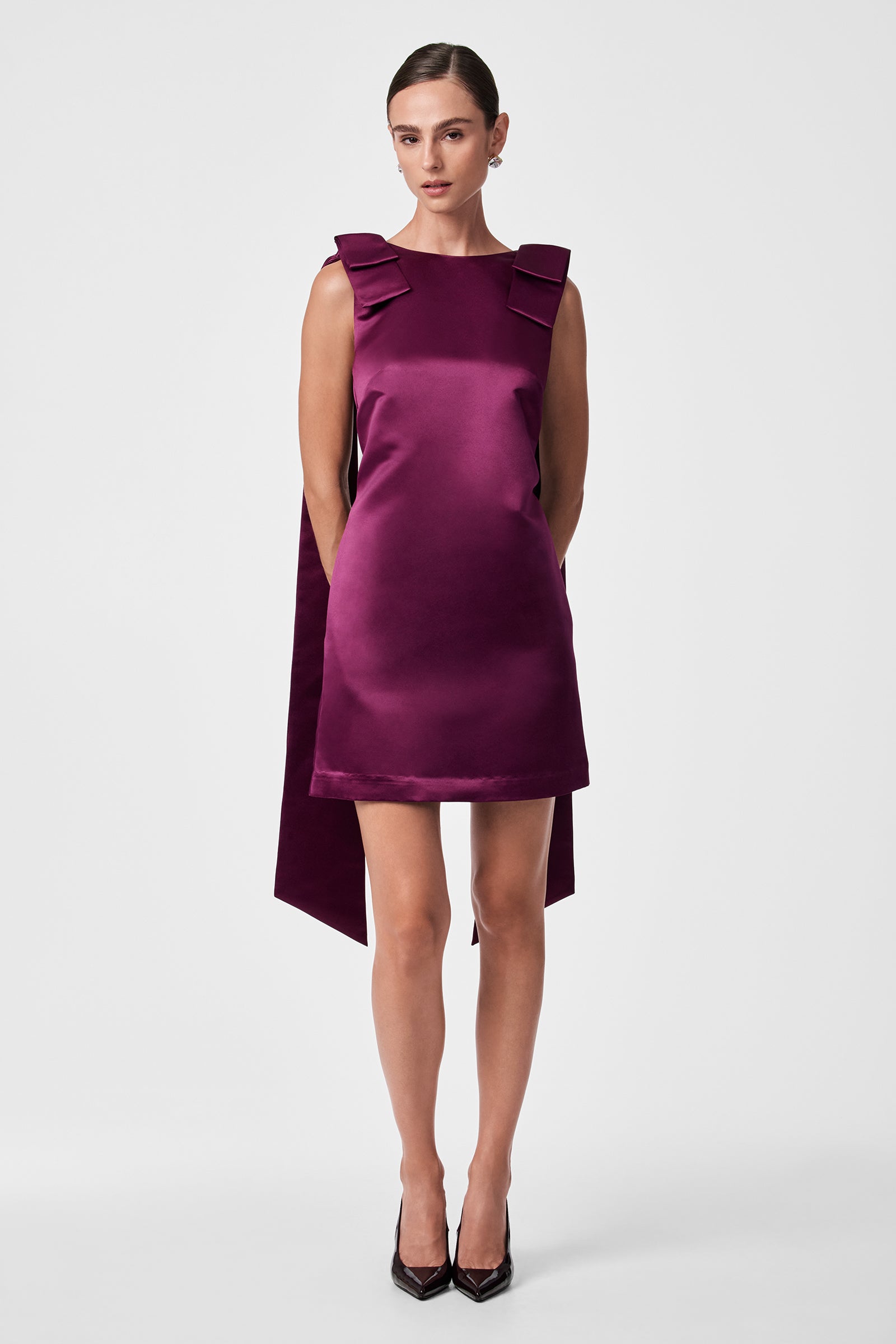 A woman wears the Priscilla Dress—a sleeveless, knee-length, shiny purple piece with large shoulder bows and a long back panel. Her hair is tied back, arms are relaxed, and she pairs it with black pointed heels against a plain backdrop.