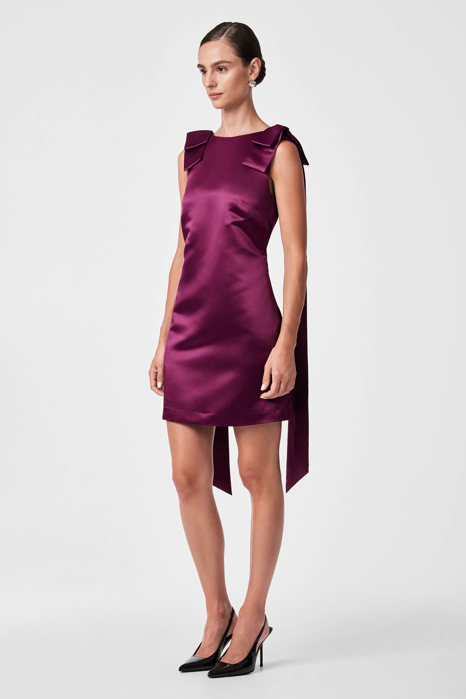 A woman in the Priscilla Dress—a sleeveless, knee-length deep purple satin dress with large shoulder bows—and black pointed heels stands against a plain light background.