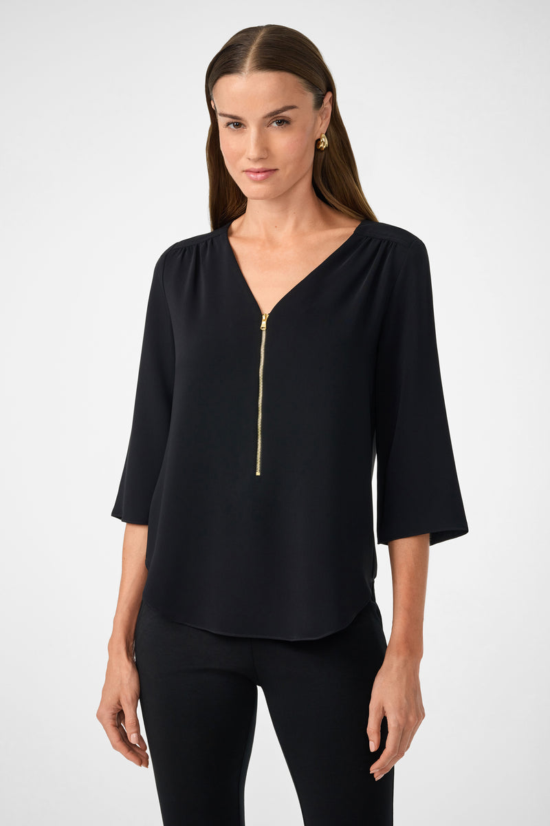 A woman with long brown hair models the Tamarac Top in black, a three-quarter sleeve blouse with a gold front zipper, paired with black pants, against a plain light background.