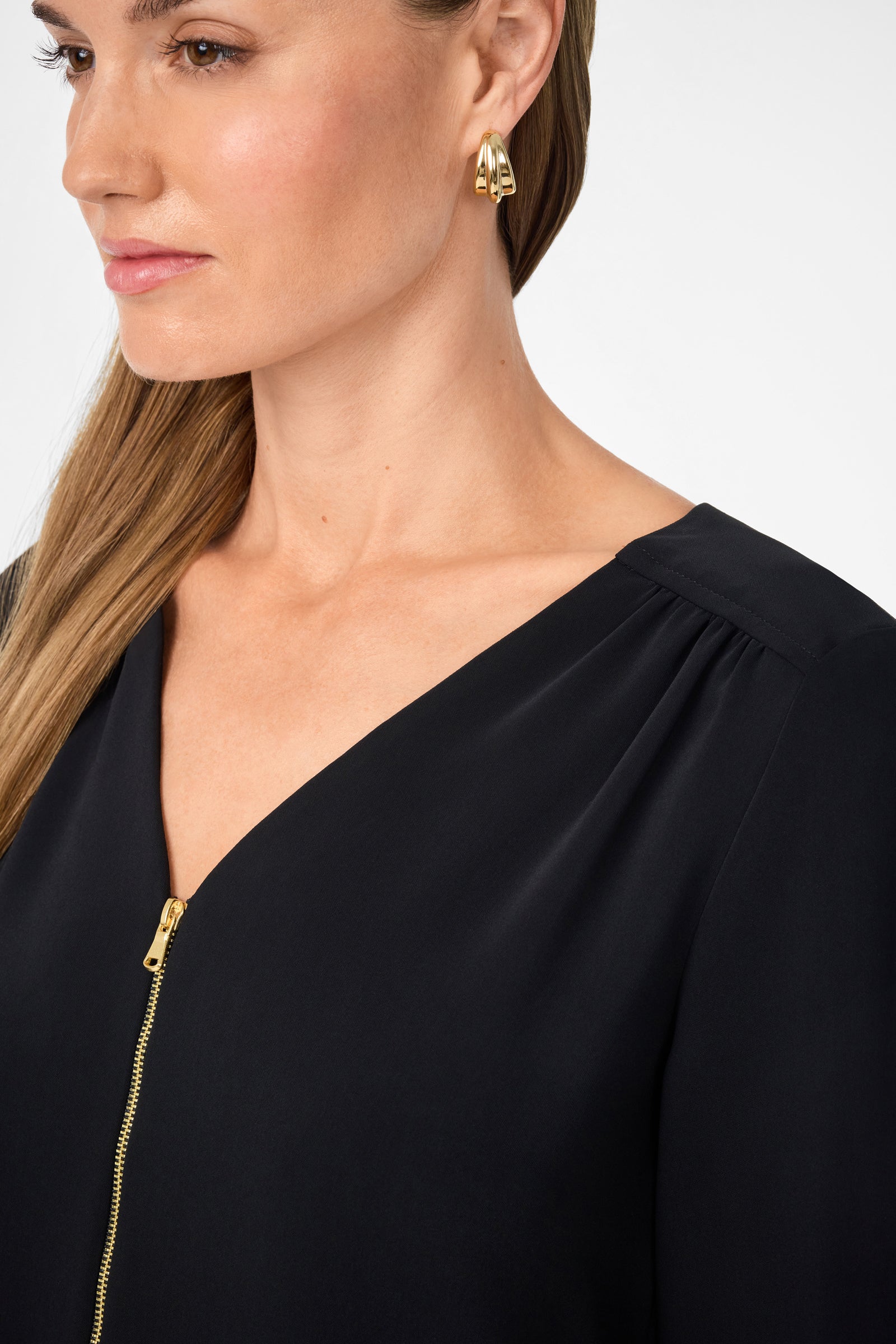 A woman wearing the Tamarac Top, a black V-neck with a gold zipper, and gold hoop earrings is shown from the shoulders up against a plain white background.