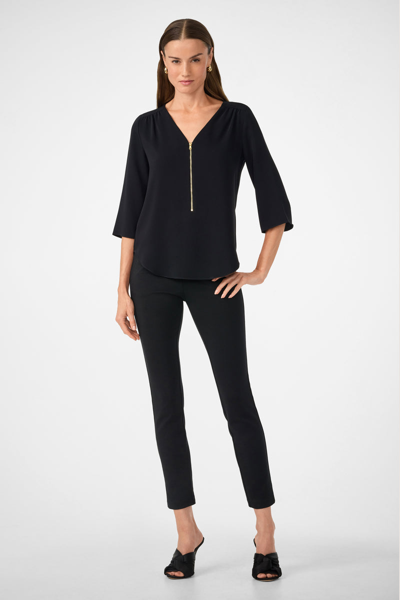 A woman with straight brown hair and a neutral expression stands against a white background, wearing the Tamarac Top—a black V-neck blouse with a front zipper—paired with black fitted pants and black open-toe heels.