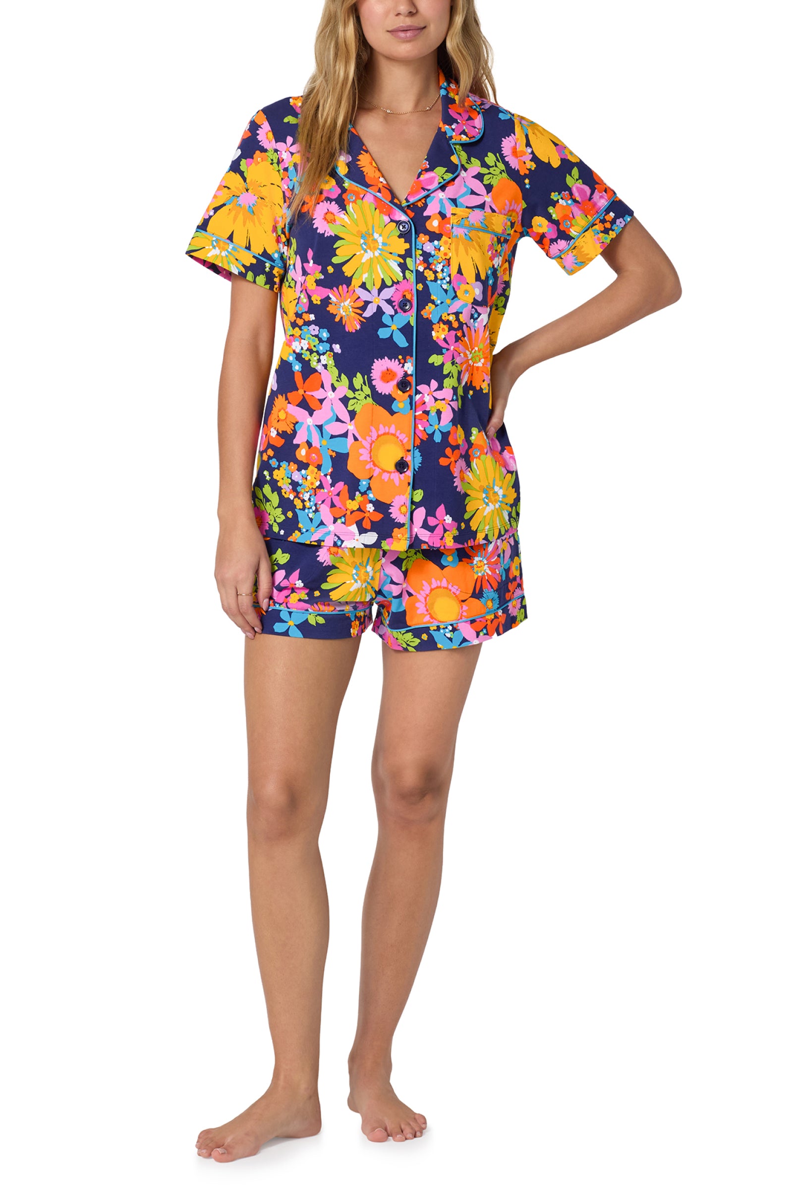 A woman stands barefoot in the Shorty PJ Set - Bay St Bloom: a short-sleeve, button-up pajama set with matching shorts, showcasing a bold, colorful floral pattern on a dark background.