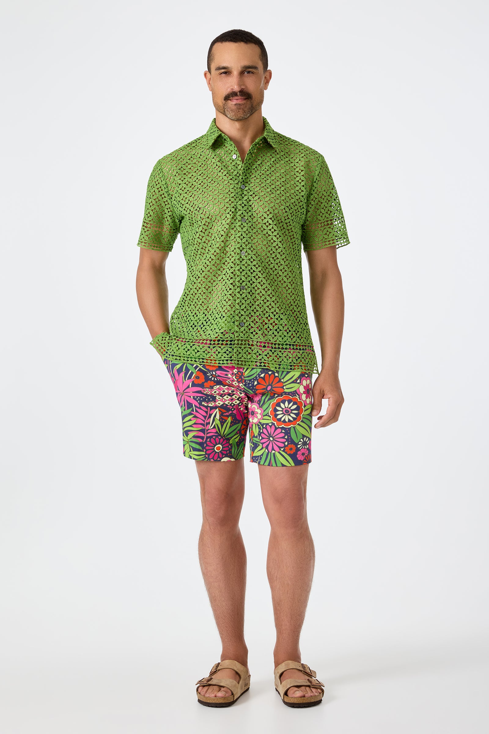 A man with short hair and a mustache stands against a white background wearing the green Belmont Shore Shirt with a perforated pattern, bright floral shorts in pink and green, and tan sandals.