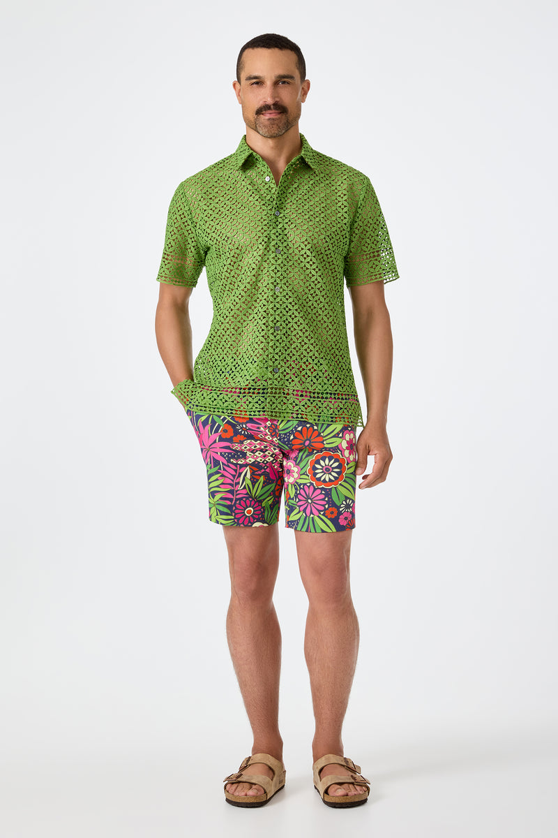 A man with short hair and a mustache stands against a white background wearing the green Belmont Shore Shirt with a perforated pattern, bright floral shorts in pink and green, and tan sandals.