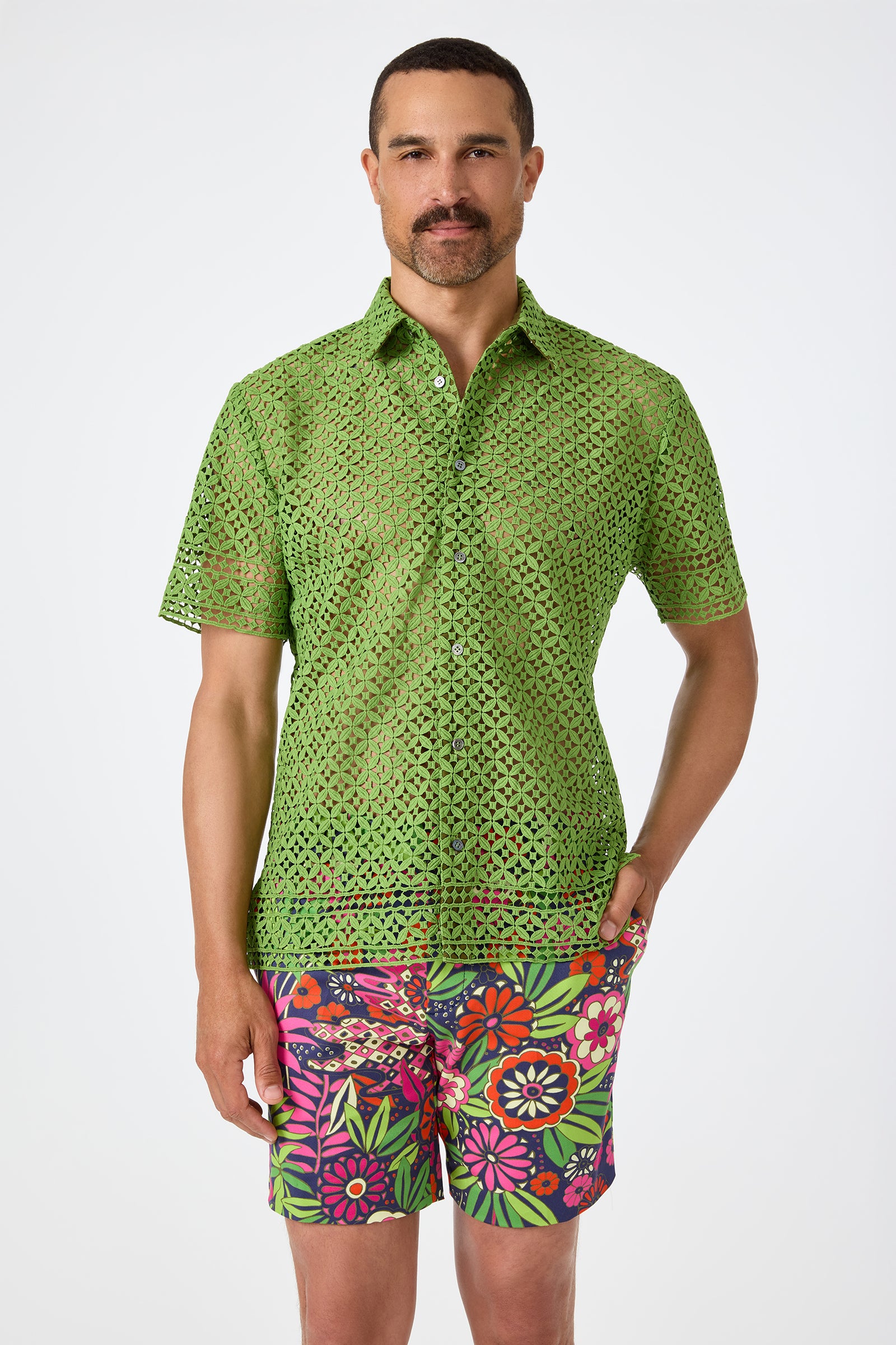 A man with short dark hair and a mustache wears the Belmont Shore Shirt in Sogen Green, a short-sleeved, perforated button-up, paired with colorful floral shorts against a plain white background.