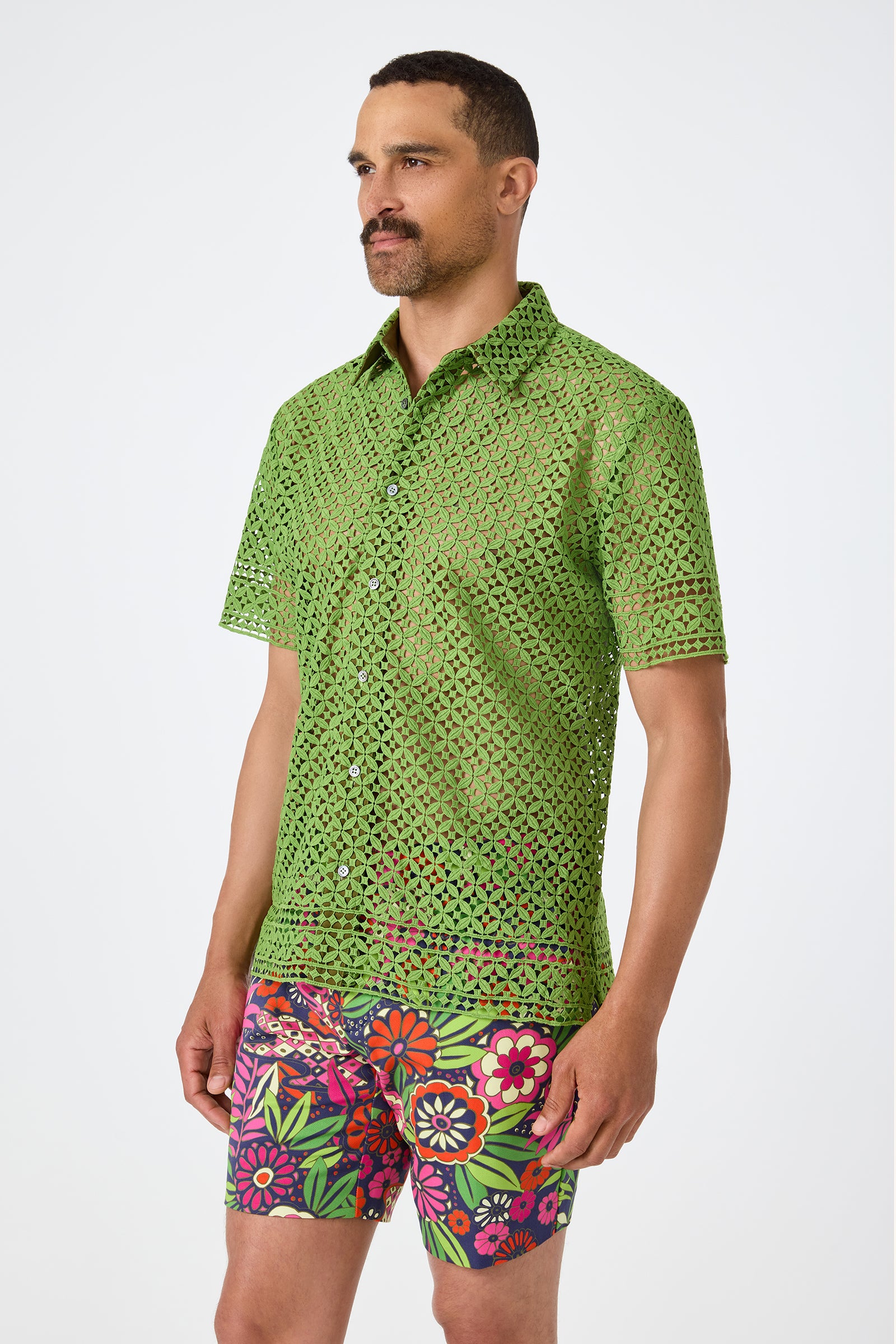 A man with short hair and a mustache wears the Belmont Shore Shirt, a sheer green short-sleeve button-up with a geometric cutout pattern, paired with colorful floral shorts. He stands against a plain white background.