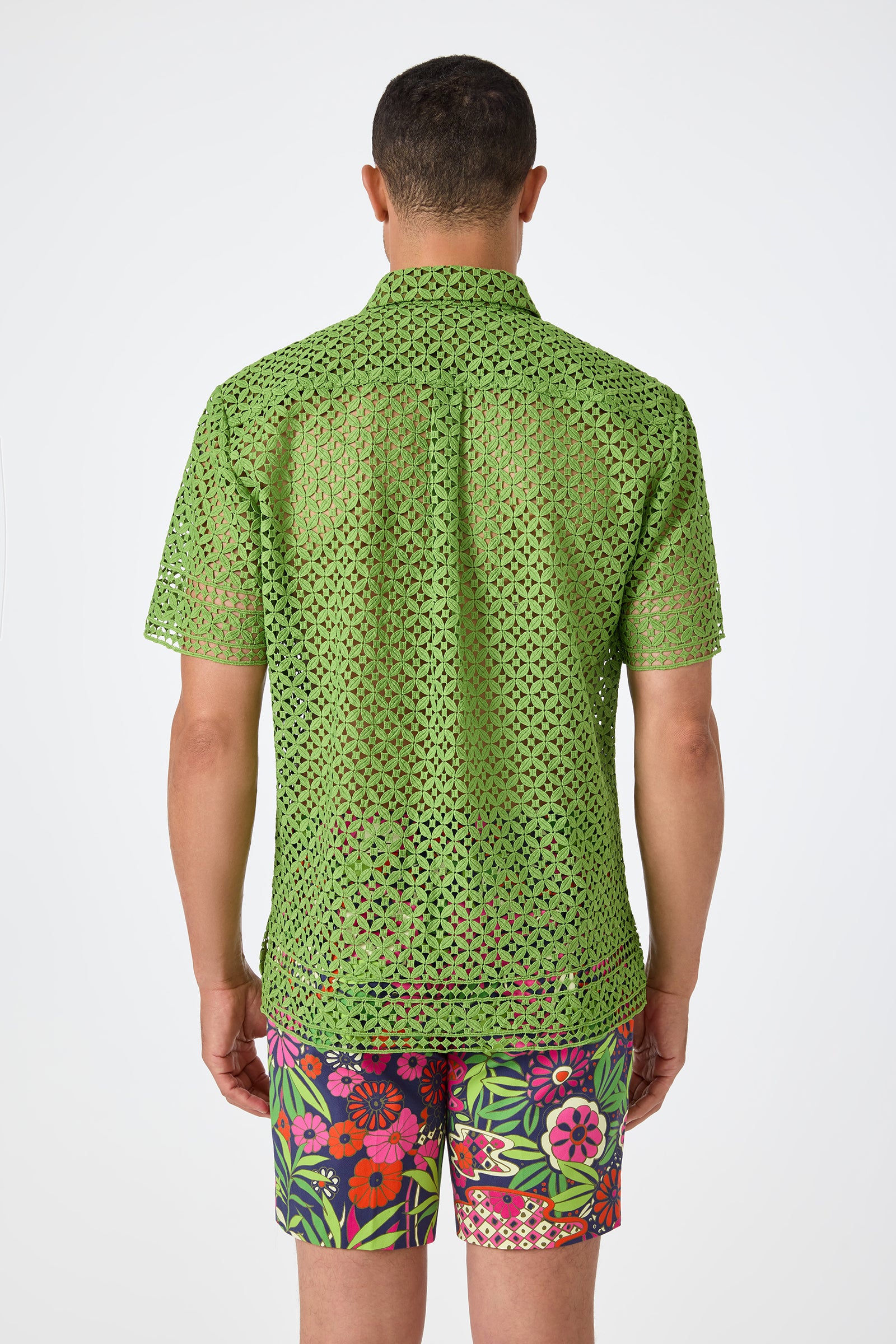 A person stands facing away, wearing the Belmont Shore Shirt—a short-sleeve green eyelet top—and brightly colored floral shorts with pink, red, and green patterns, set against a plain white background.