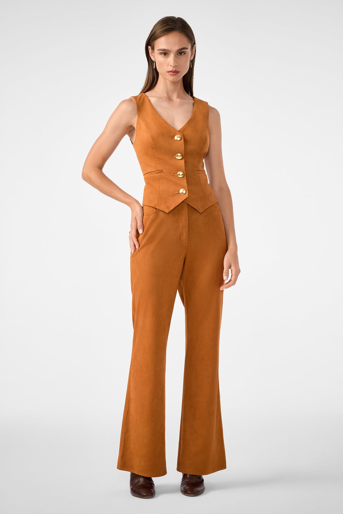 A woman faces forward wearing the Kofi 2 Vest—a sleeveless, fitted tan vest with large gold buttons—and matching high-waisted, flared pants. She has straight brown hair, brown shoes, and stands against a plain light background.