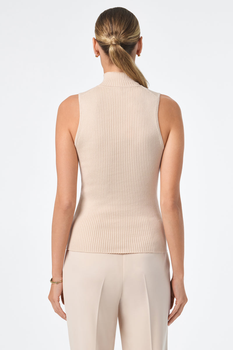 A woman with her hair in a low ponytail faces away, wearing the Westhampton Knit Pullover—a sleeveless, beige ribbed turtleneck top—paired with light pants. The background is plain white.