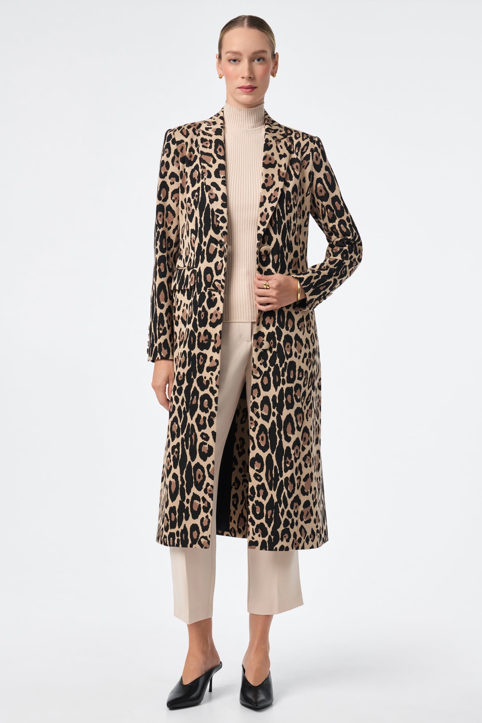 A woman stands against a white background wearing the Playbill Coat, a long leopard print piece, over a beige turtleneck sweater, cream ankle-length trousers, and black heeled shoes. Her hair is pulled back and her expression is neutral.
