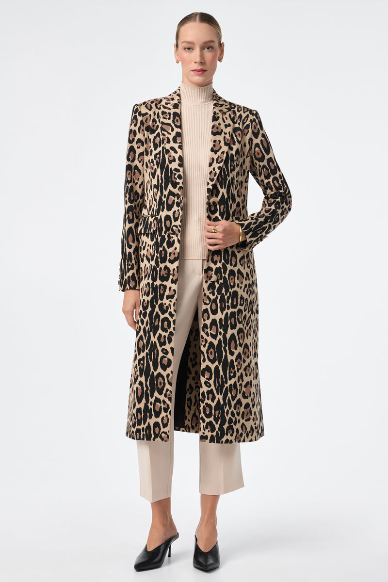A woman stands against a white background wearing the Playbill Coat, a long leopard print piece, over a beige turtleneck sweater, cream ankle-length trousers, and black heeled shoes. Her hair is pulled back and her expression is neutral.