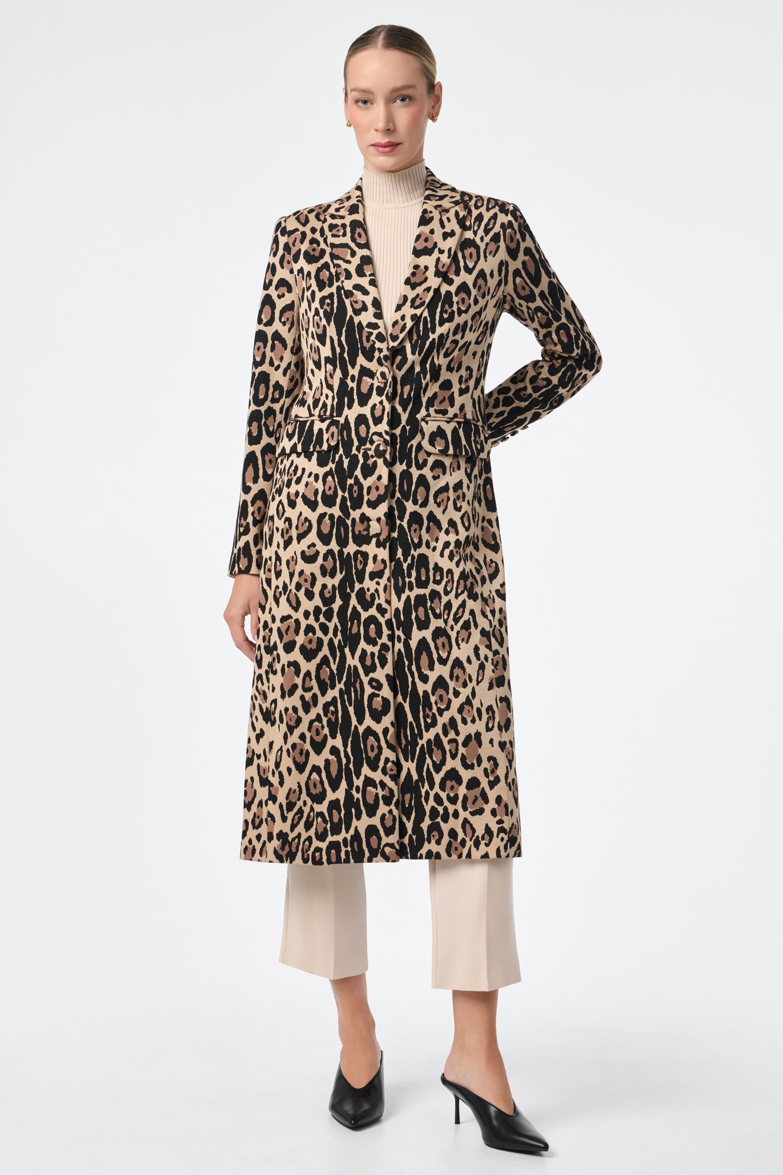 A woman stands against a white background wearing the Playbill Coat, a long beige and black leopard print style, over a light turtleneck, cream cropped pants, and black pointed-toe heels. Her hair is pulled back and her hands are at her sides.
